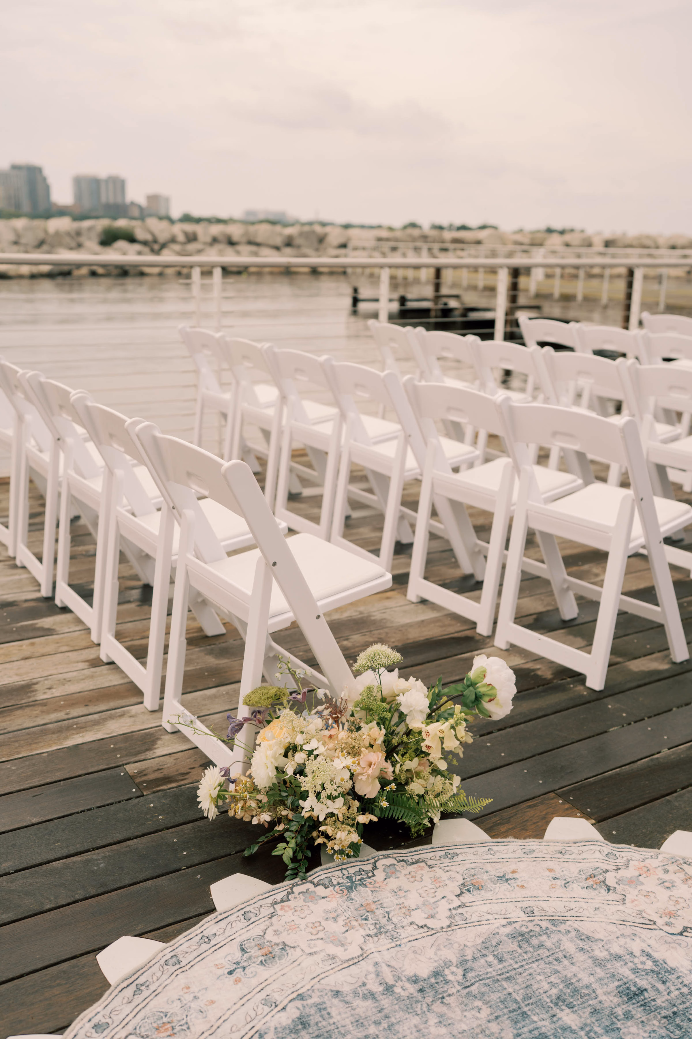Event Essentials white resin folding chairs with padded seats arranged on a dark wood deck for an outdoor ceremony, accented by a wildflower floral arrangement and a distressed blue area rug.