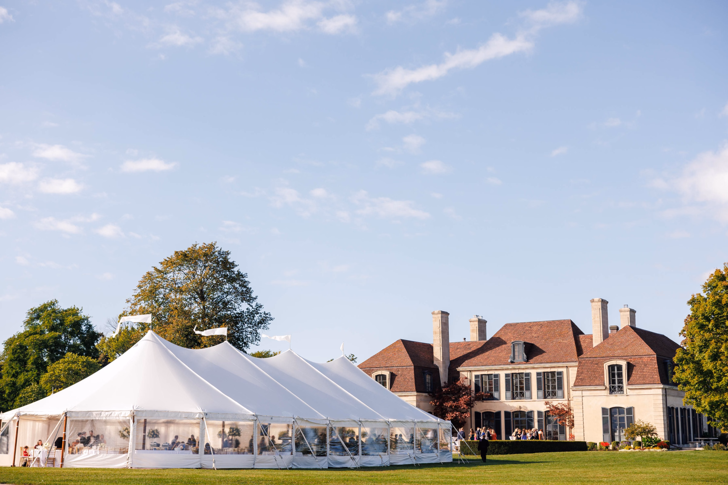A large white Event Essentials sailcloth tent with clear side walls and white flags installed on the expansive lawn of a luxury historic estate for an outdoor wedding reception.