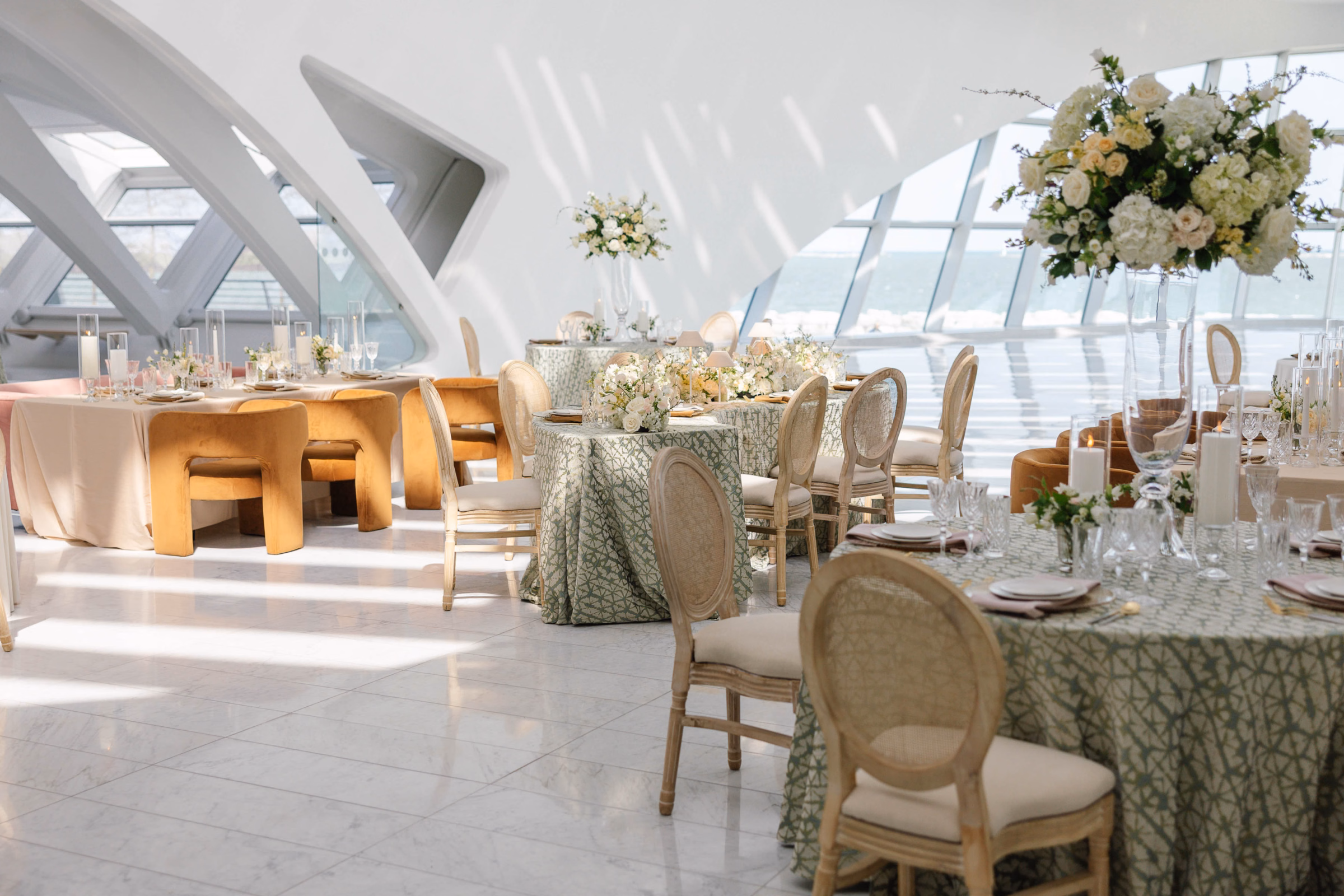 An Event Essentials luxury reception setup featuring sage green geometric patterned tablecloths, round-back cane Louis chairs, ocher velvet accent chairs, and tall floral centerpieces in a bright modern venue.