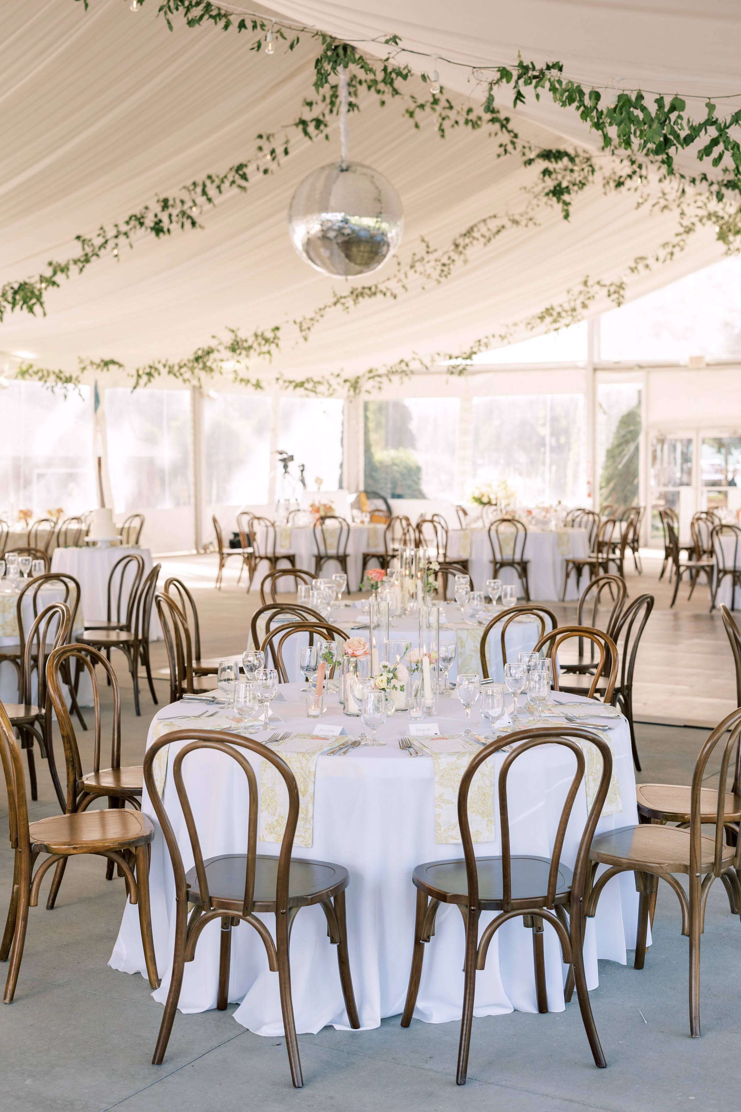 Interior of an Event Essentials wedding tent featuring round tables with white linens, wooden bentwood chairs, a large hanging silver disco ball, and greenery garlands draping the fabric-lined ceiling.