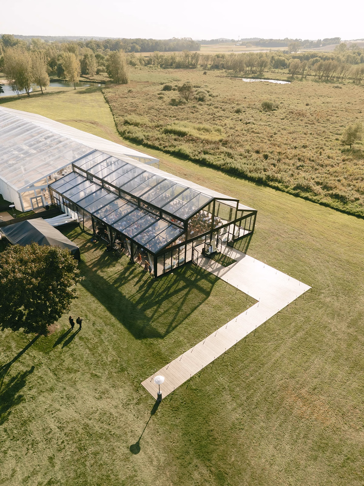 High-angle aerial view of an Event Essentials glass-walled structure tent with a black metal frame and clear-top roof, featuring a custom wooden boardwalk walkway connecting to a large white reception tent on a lawn.