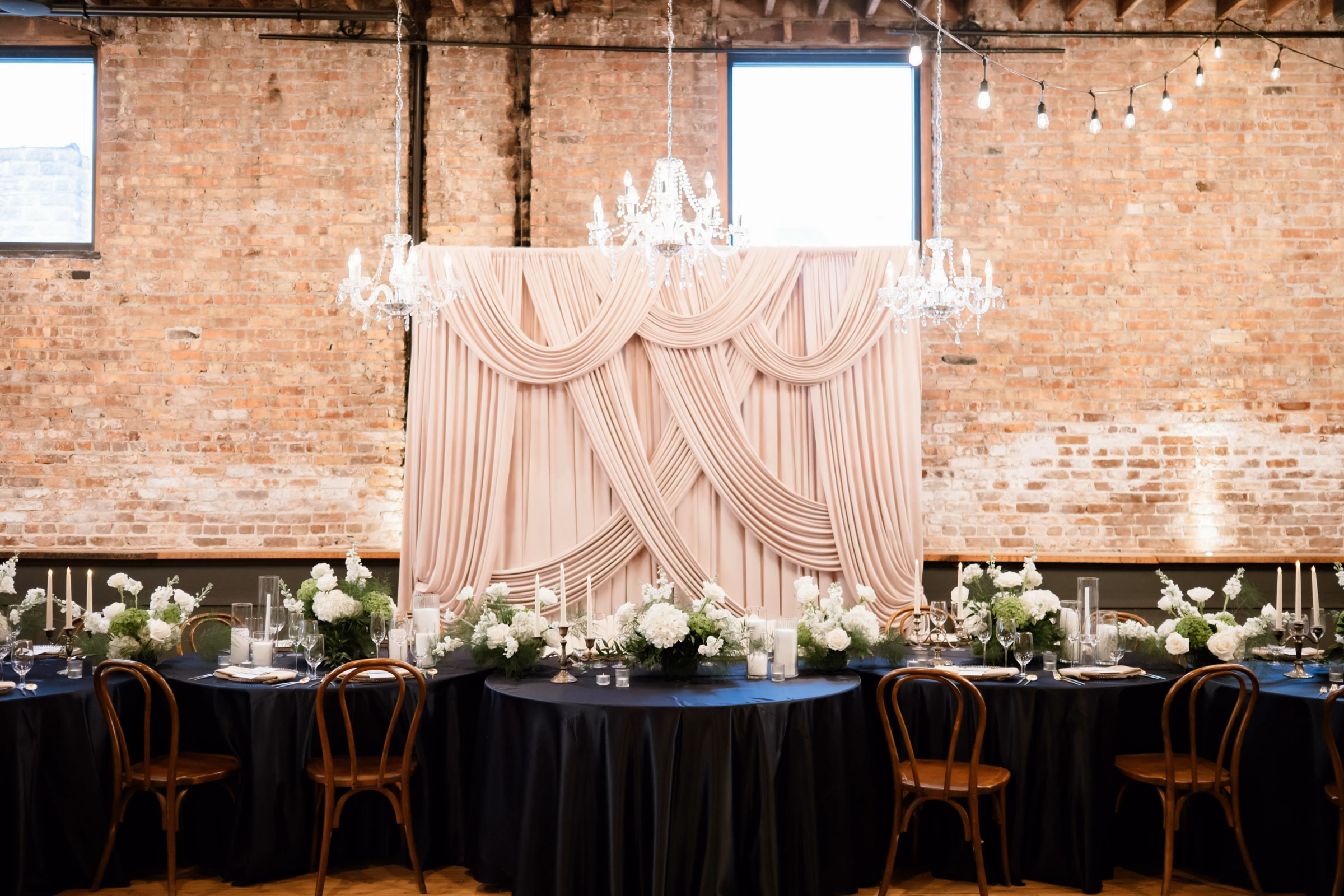 An Event Essentials indoor wedding setup featuring a head table with black linens and wooden bentwood chairs, backed by an ornate blush fabric drapery backdrop and three hanging crystal chandeliers against an exposed brick wall.