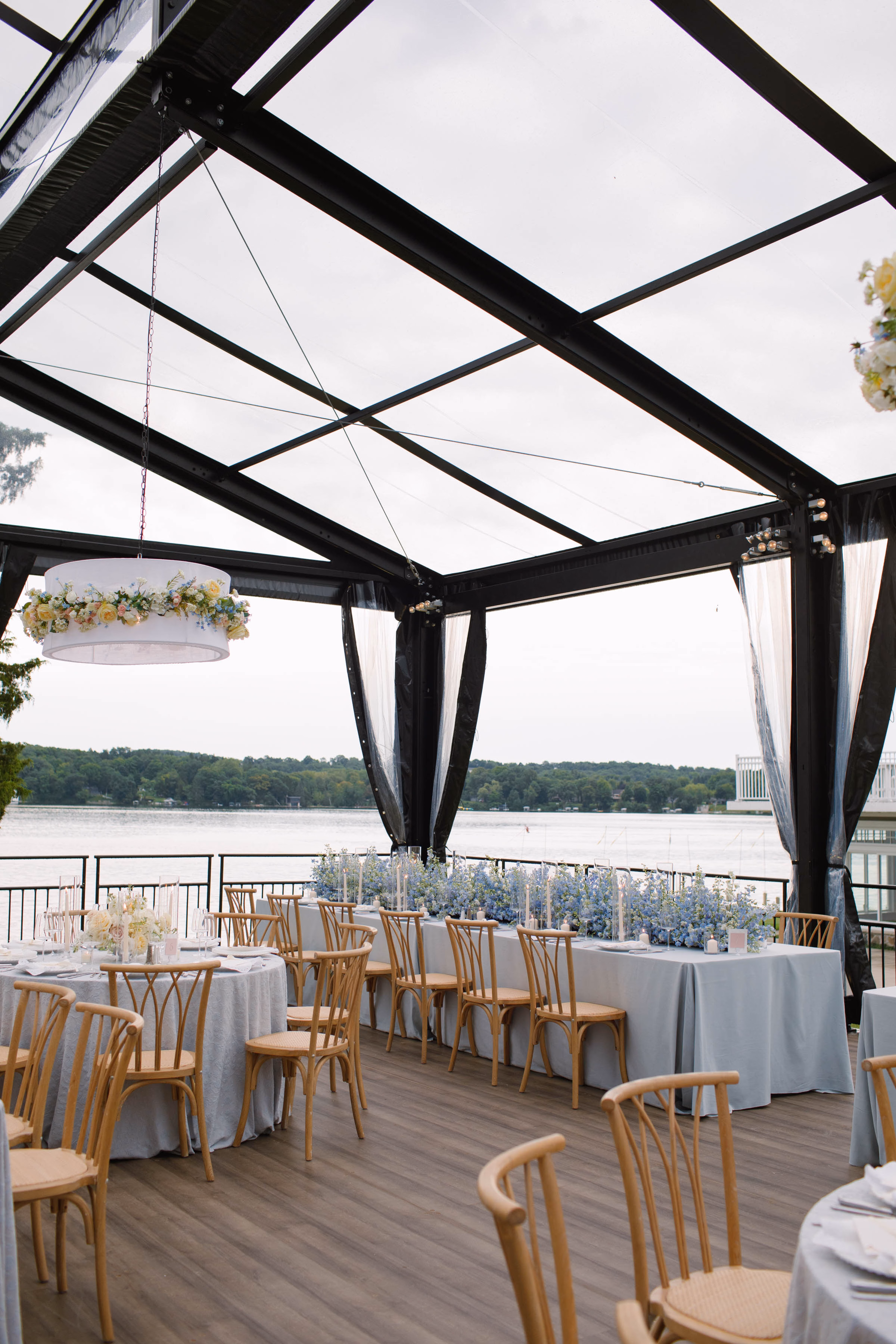 Interior of an Event Essentials clear-top glass structure tent with a black metal frame, featuring light gray wooden flooring, natural wood chairs, light blue linens, and a custom hanging drum pendant light wrapped in floral garlands.