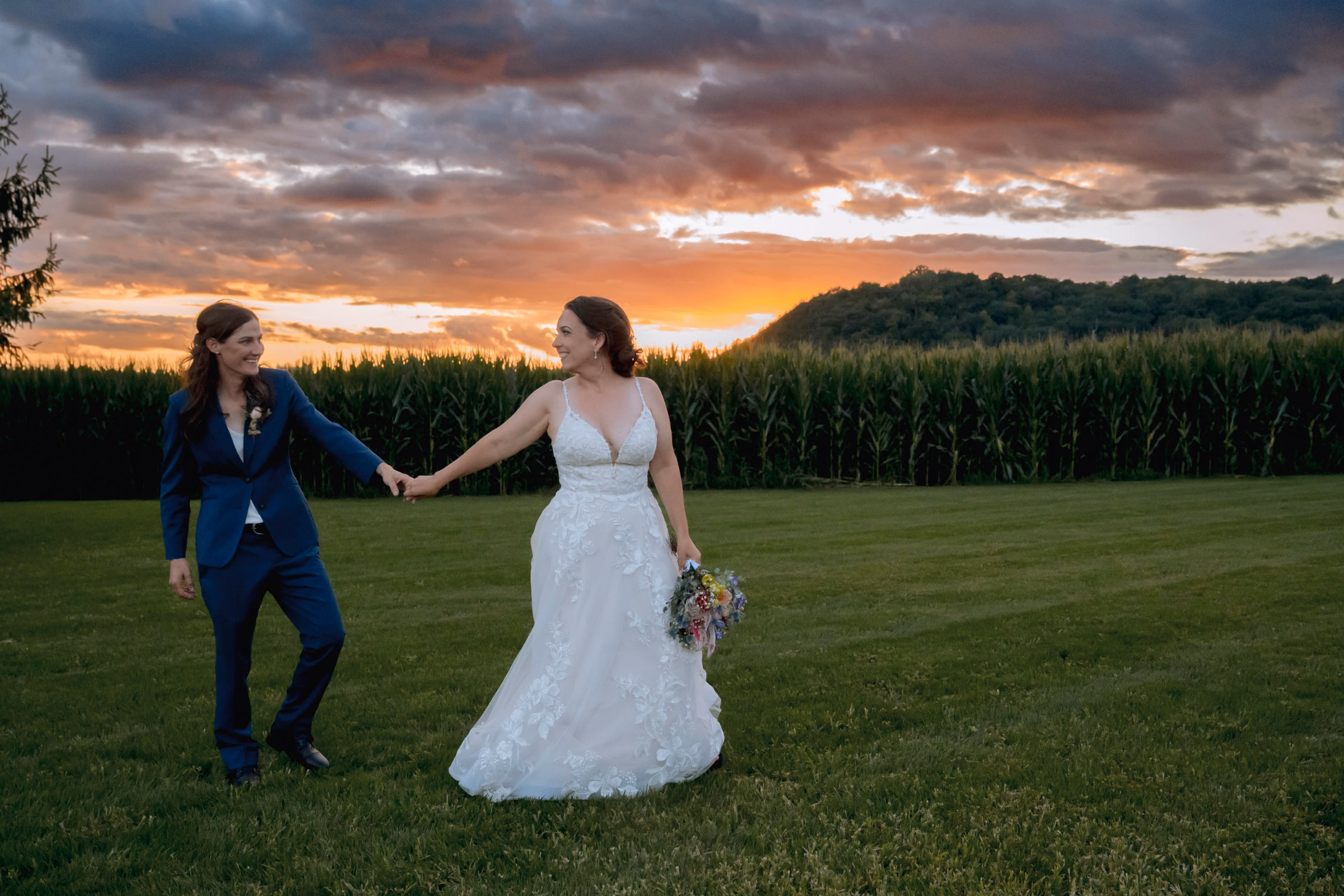 Two brides holding hands and smiling at each other in a grassy field in front of a cornfield during a dramatic sunset, by Reminisce Photography