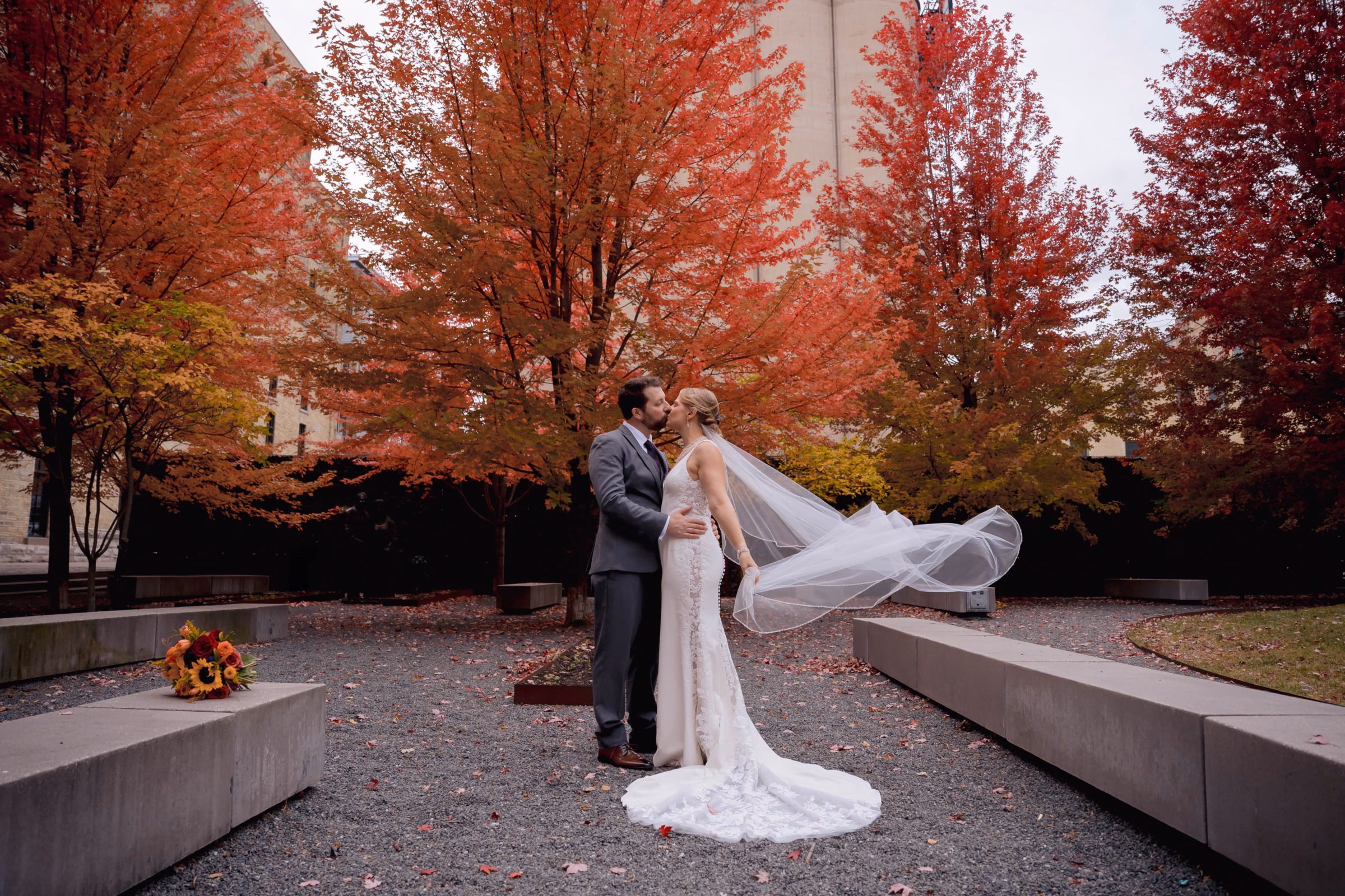 A bride and groom kissing surrounded by vibrant red autumn trees as the bride's long veil floats behind her, by Reminisce Photography