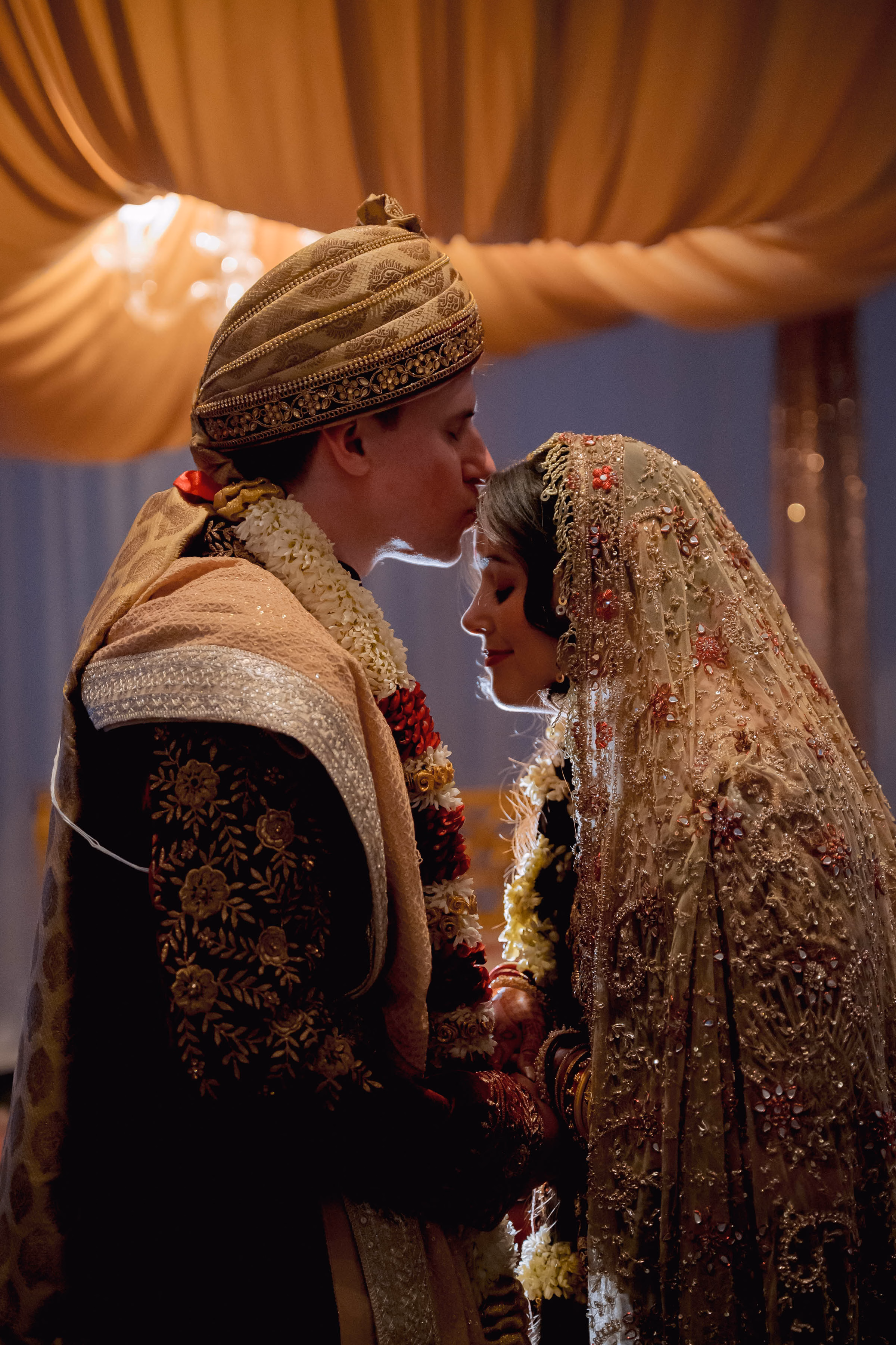 An intimate, backlit wedding portrait of a groom kissing his bride's forehead during a traditional South Asian wedding ceremony, by Reminisce Photography