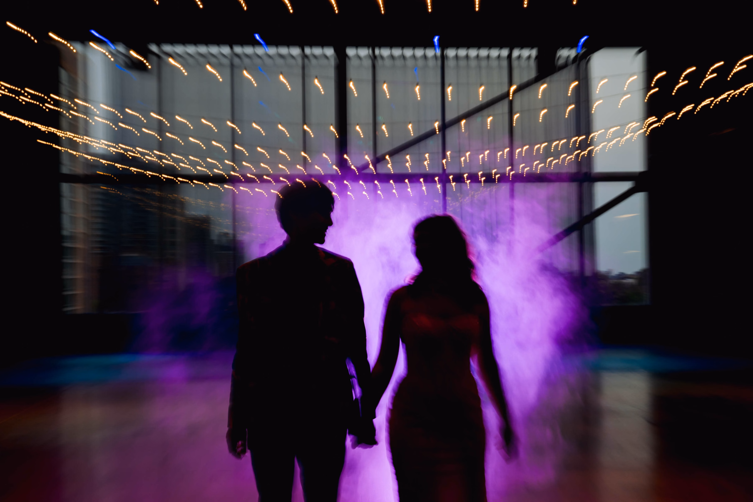 A creative motion-blur wedding portrait of a bride and groom's silhouettes against vibrant purple reception lighting and string lights, by Reminisce Photography