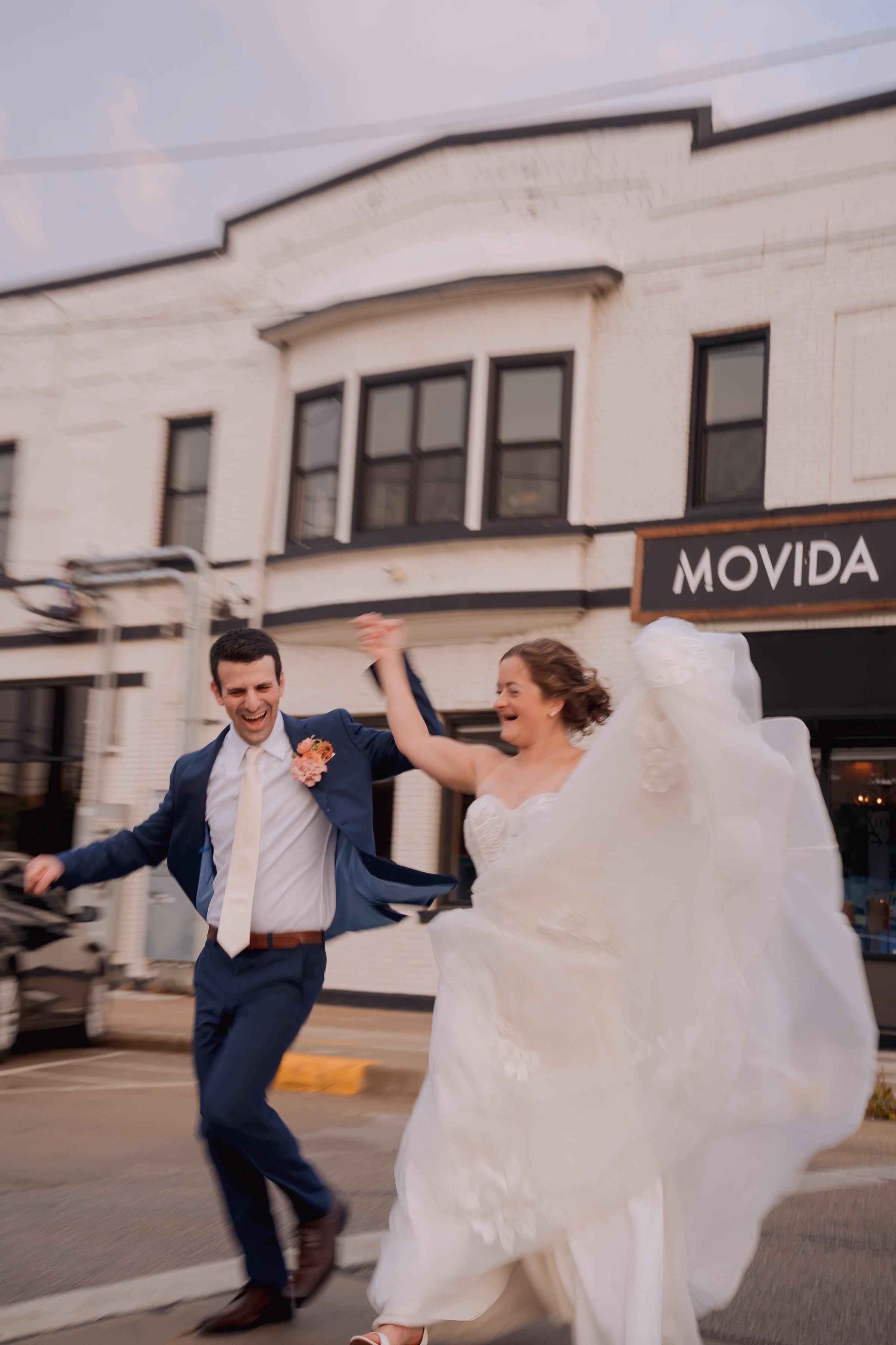 A joyful bride and groom laughing and dancing down a city street together past an urban building, by Reminisce Photography