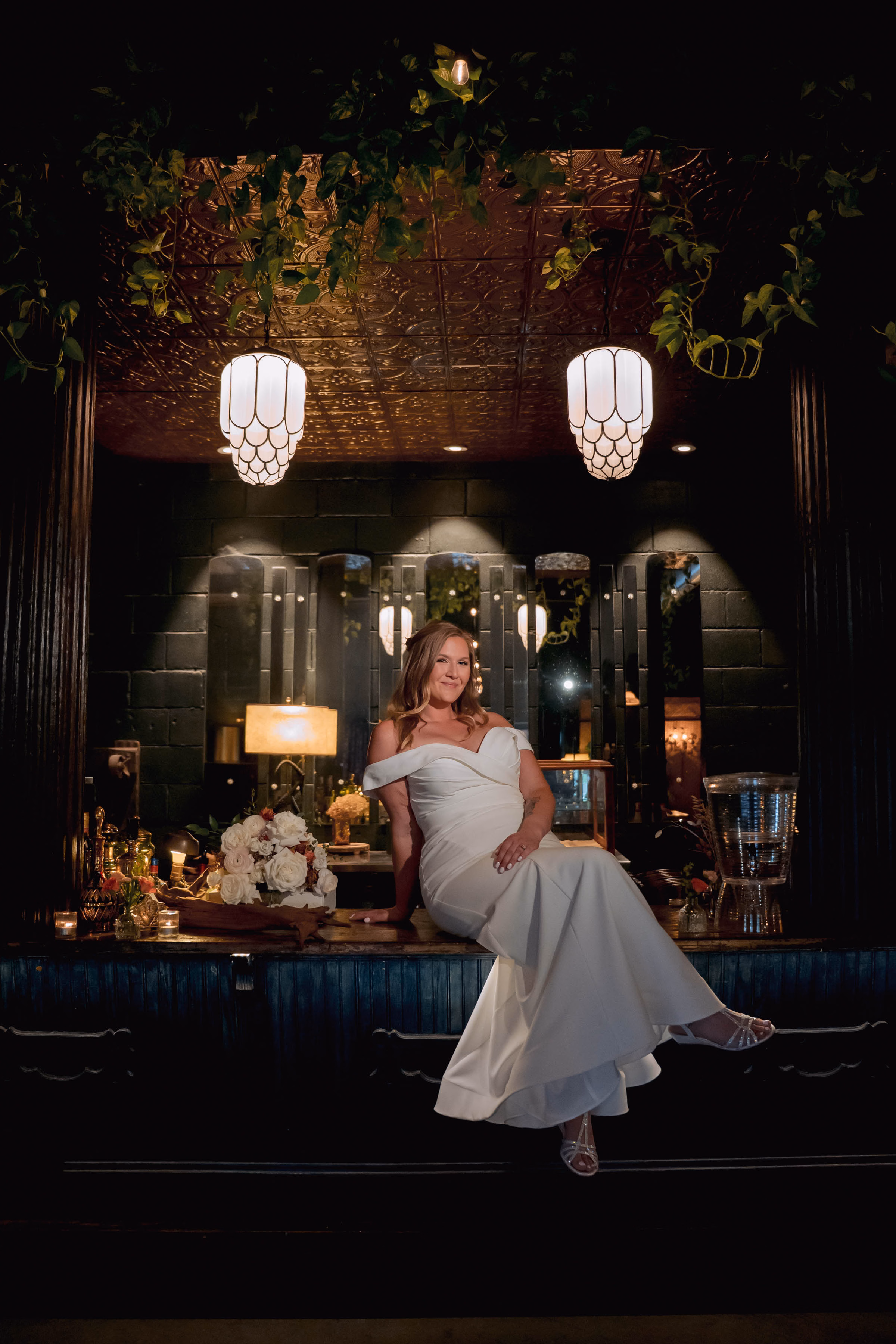 A moody, elegant bridal portrait of a bride in an off-the-shoulder gown sitting on a dark bar under warm, hanging light fixtures, by Reminisce Photography
