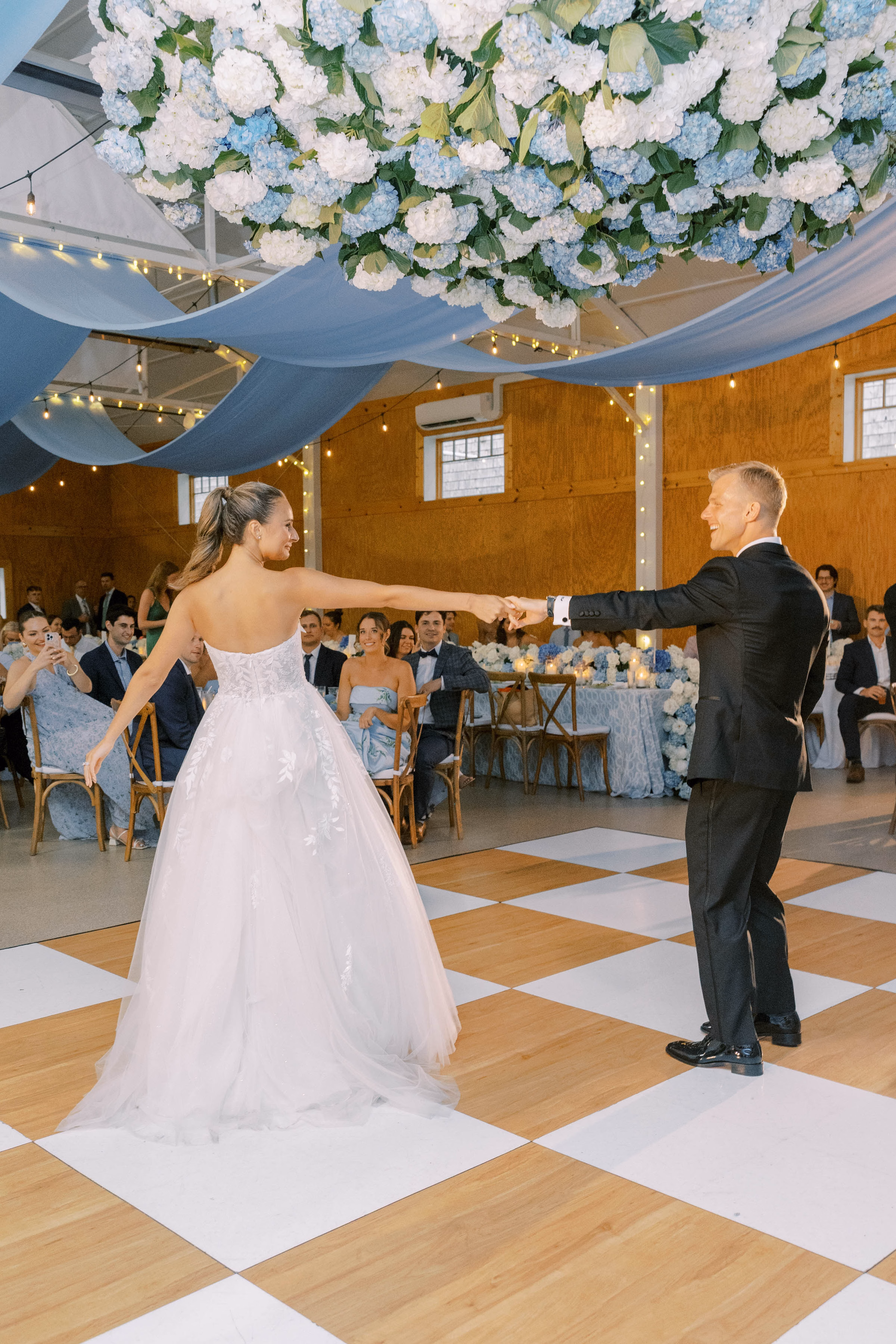 Luxury wedding reception rentals in Milwaukee, WI, by Tablescapes Event Rentals, featuring a wood and white checkered dance floor, cross-back wood chairs, and light blue ceiling fabric draping in a rustic modern venue.