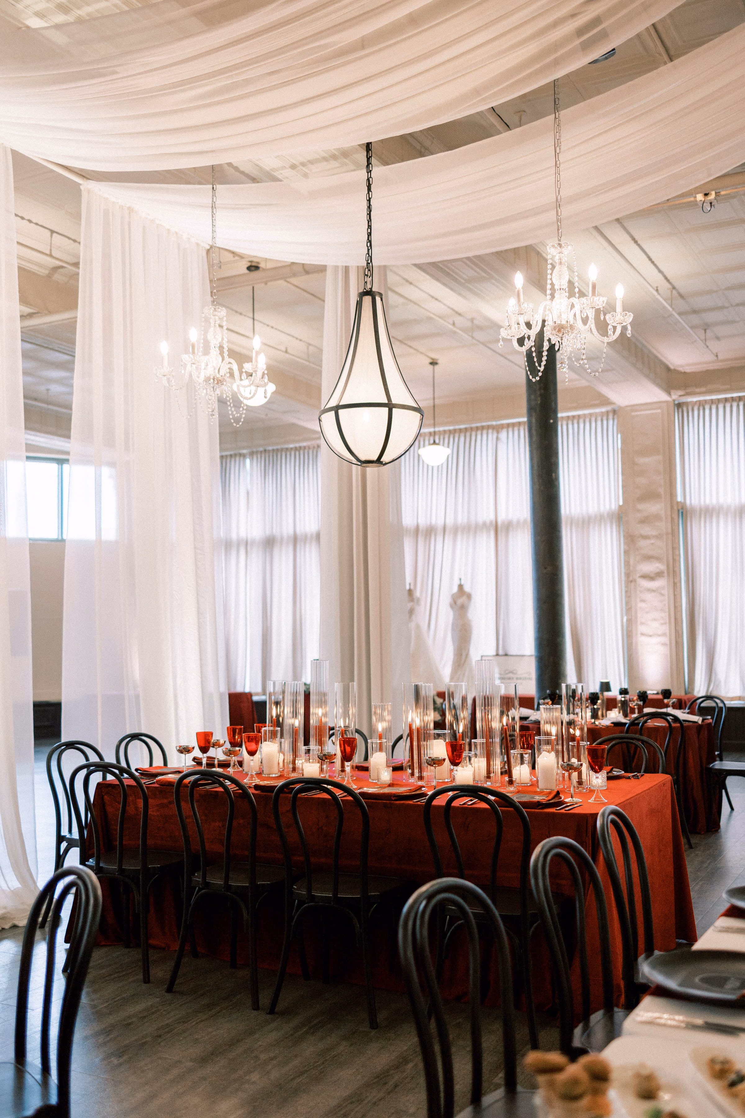 Industrial chic wedding reception tablescape rentals in Milwaukee, WI, by Tablescapes Event Rentals, featuring a red velvet tablecloth, black bentwood chairs, pillar candles in glass cylinders, and crystal chandeliers with white ceiling drapery at Pritzlaff Events.