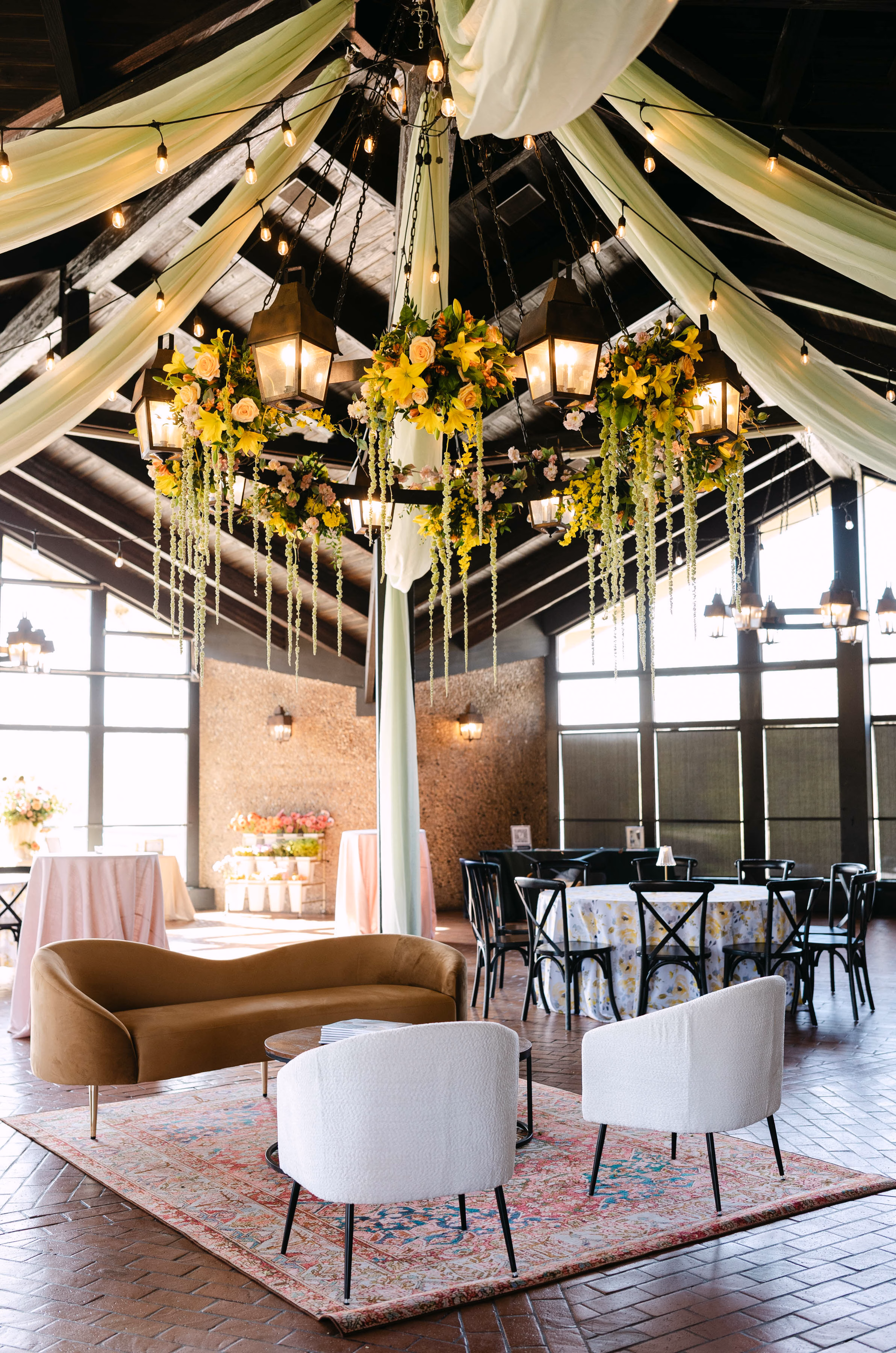Luxury lounge furniture and reception rentals in Milwaukee, WI, by Tablescapes Event Rentals, showcasing a curved mustard velvet sofa, modern white accent chairs, a vintage area rug, black cross-back chairs, and sage green ceiling drapery in a rustic pavilion.