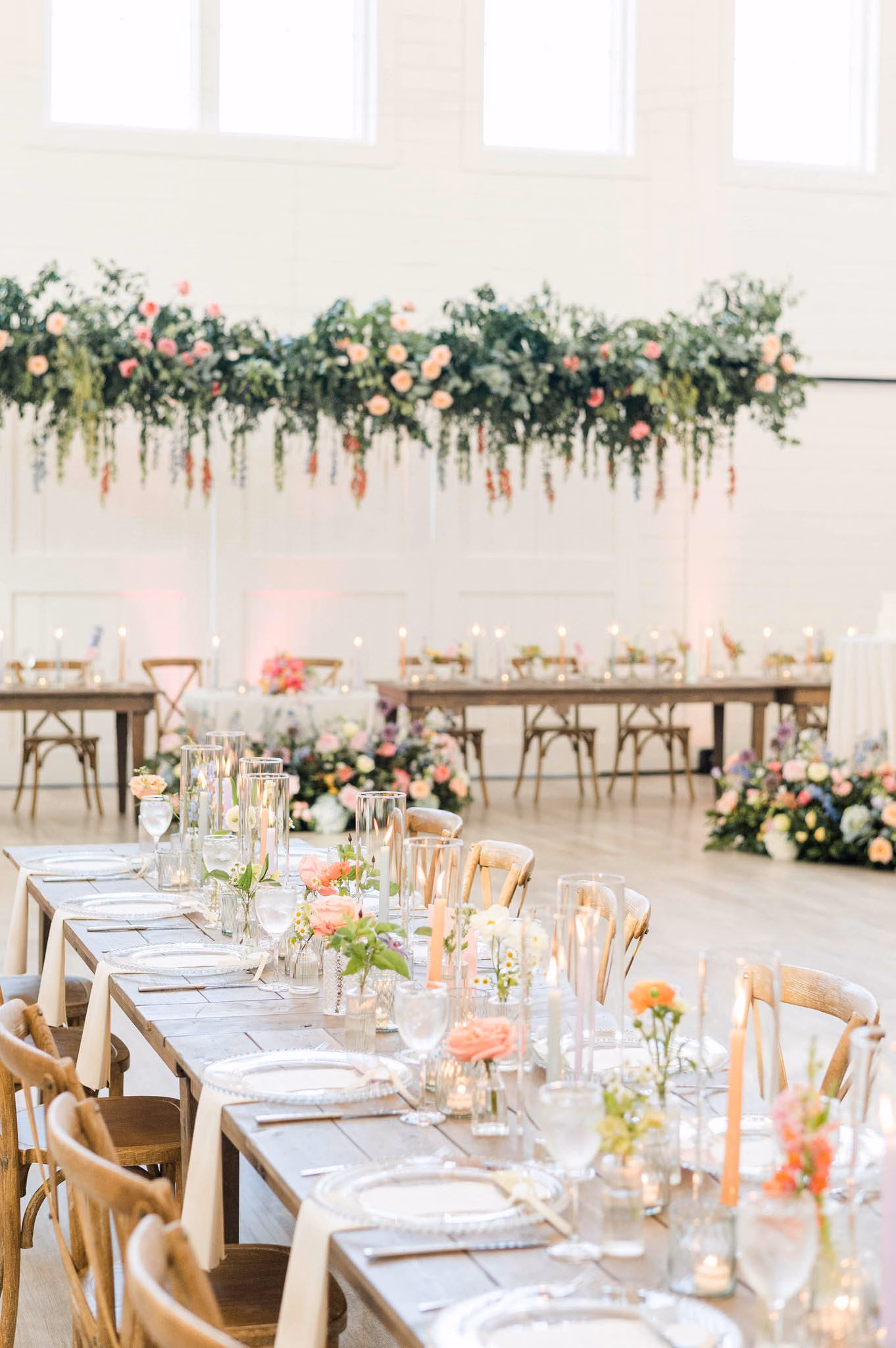 Luxury wedding tablescape and furniture rentals in Milwaukee, WI, by Tablescapes Event Rentals, featuring rustic wood farm tables, cross-back chairs, clear glass charger plates, elegant stemware, and glowing candle holders in a modern white venue.