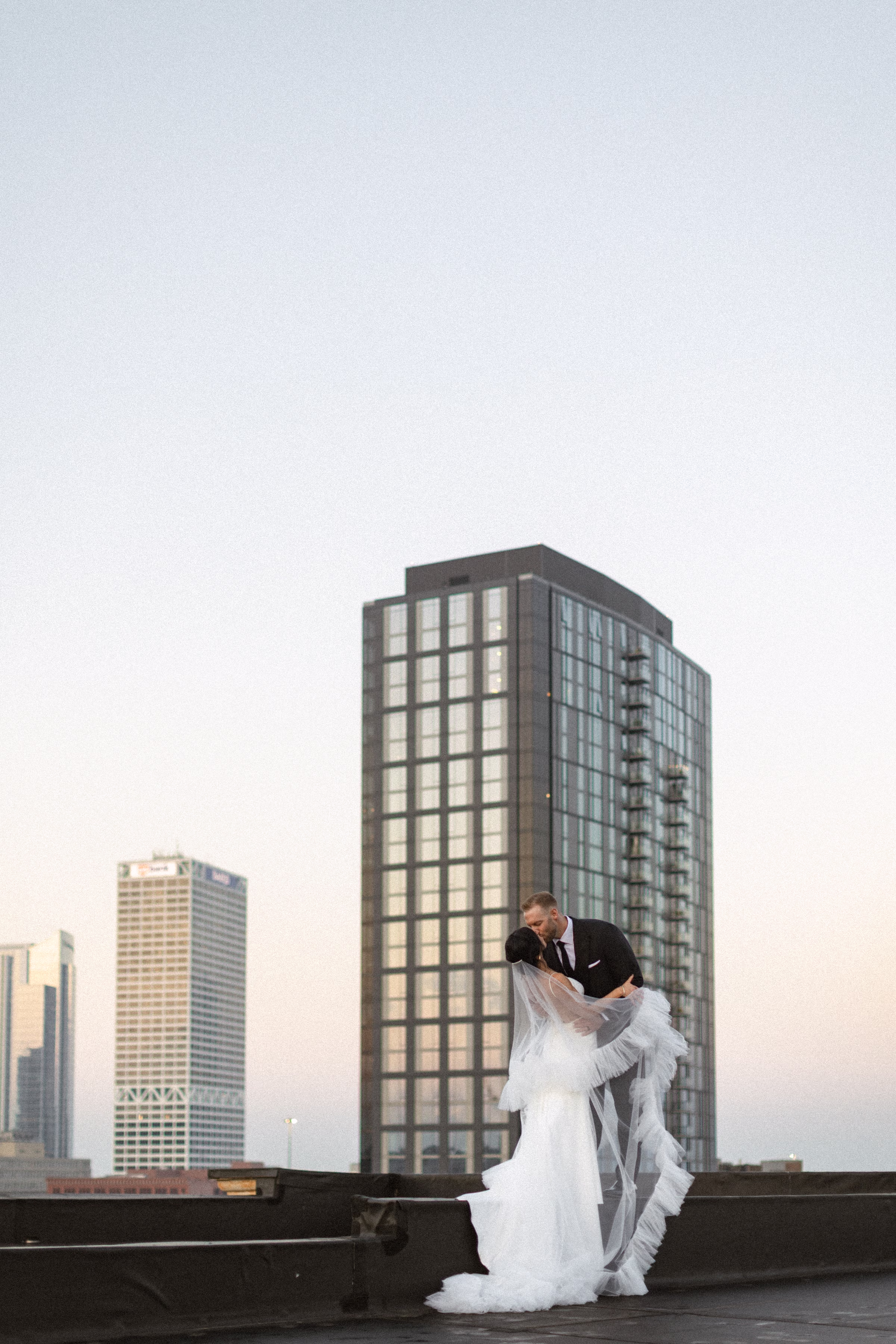 a newlywed couple shares a kiss, set against the Milwaukee skyline at sunset