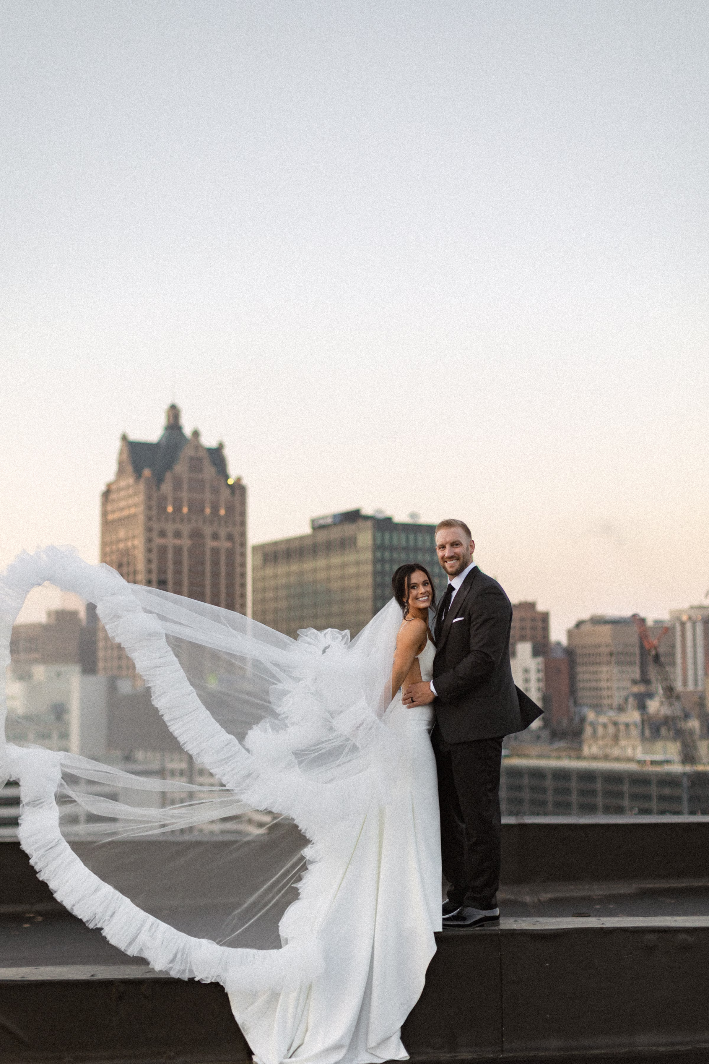 A newlywed couple smiles, set against the Milwaukee skyline at sunset. The bride has a statement, tiered-tulle veil that is blowing in the wind