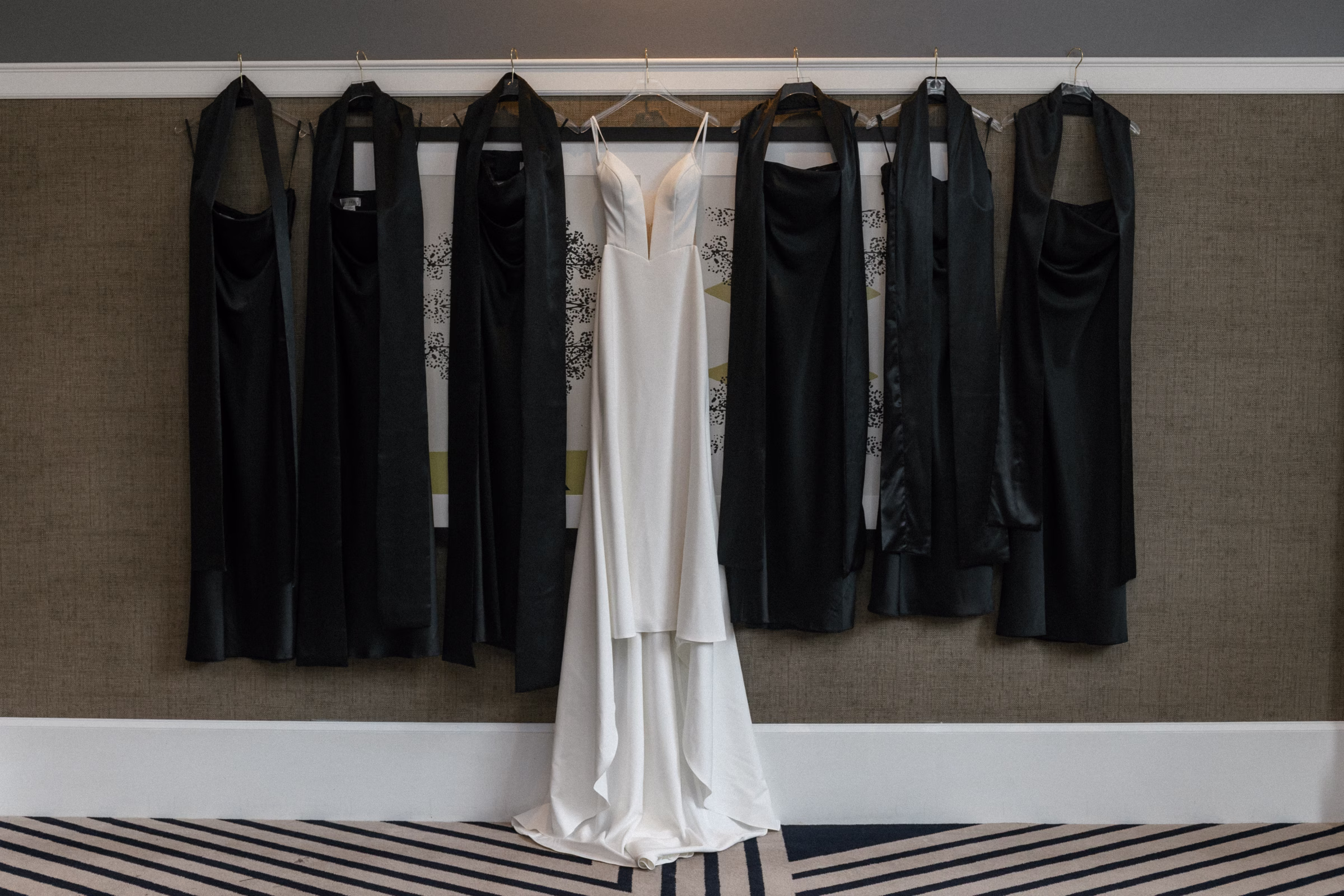 a bridal gown from Vow'd Weddings is hung up surrounded by black bridesmaids dresses from Dessy Group After Six