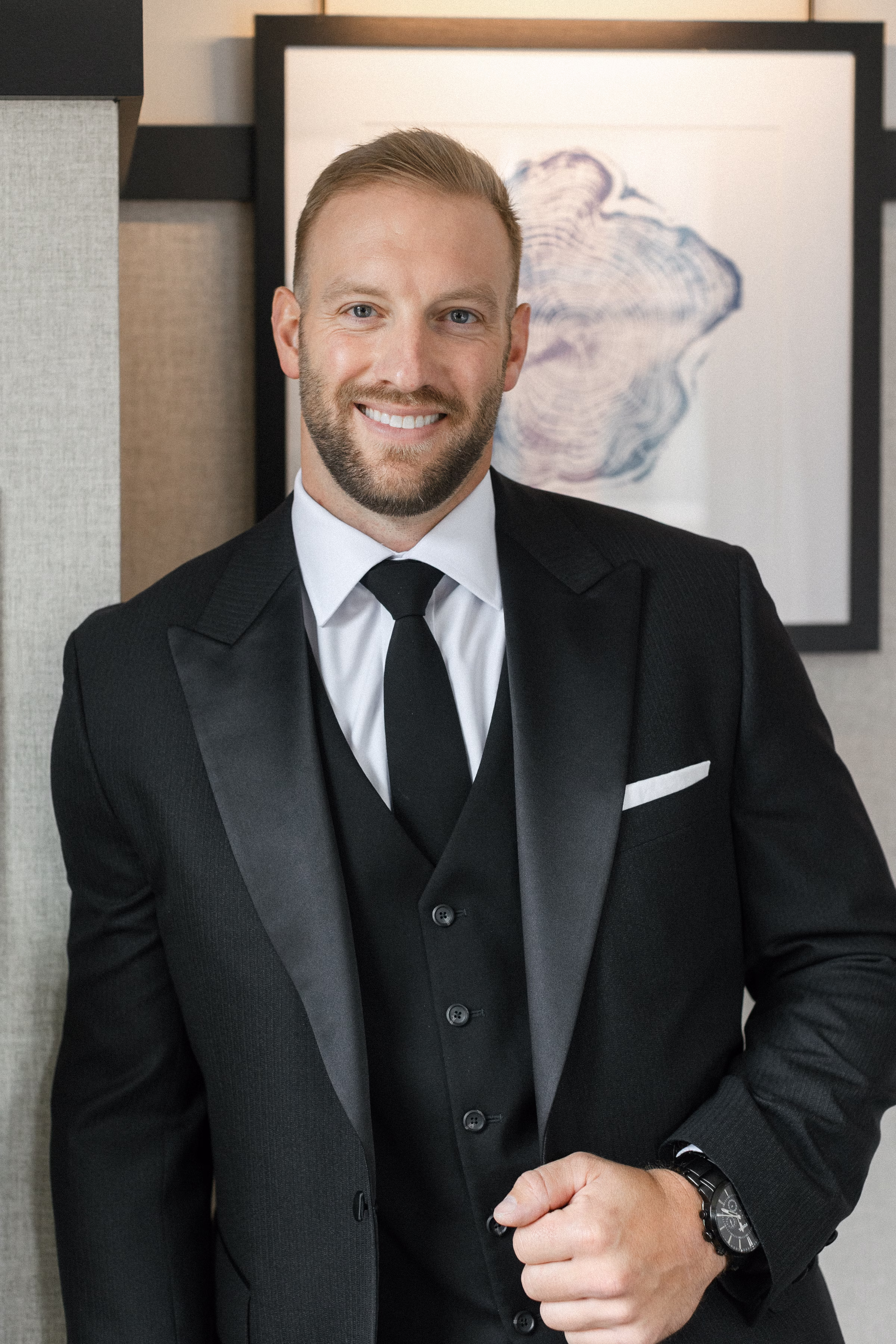a groom stands in a black tuxedo with black vest, black tie, white dress shirt, and white pocket square