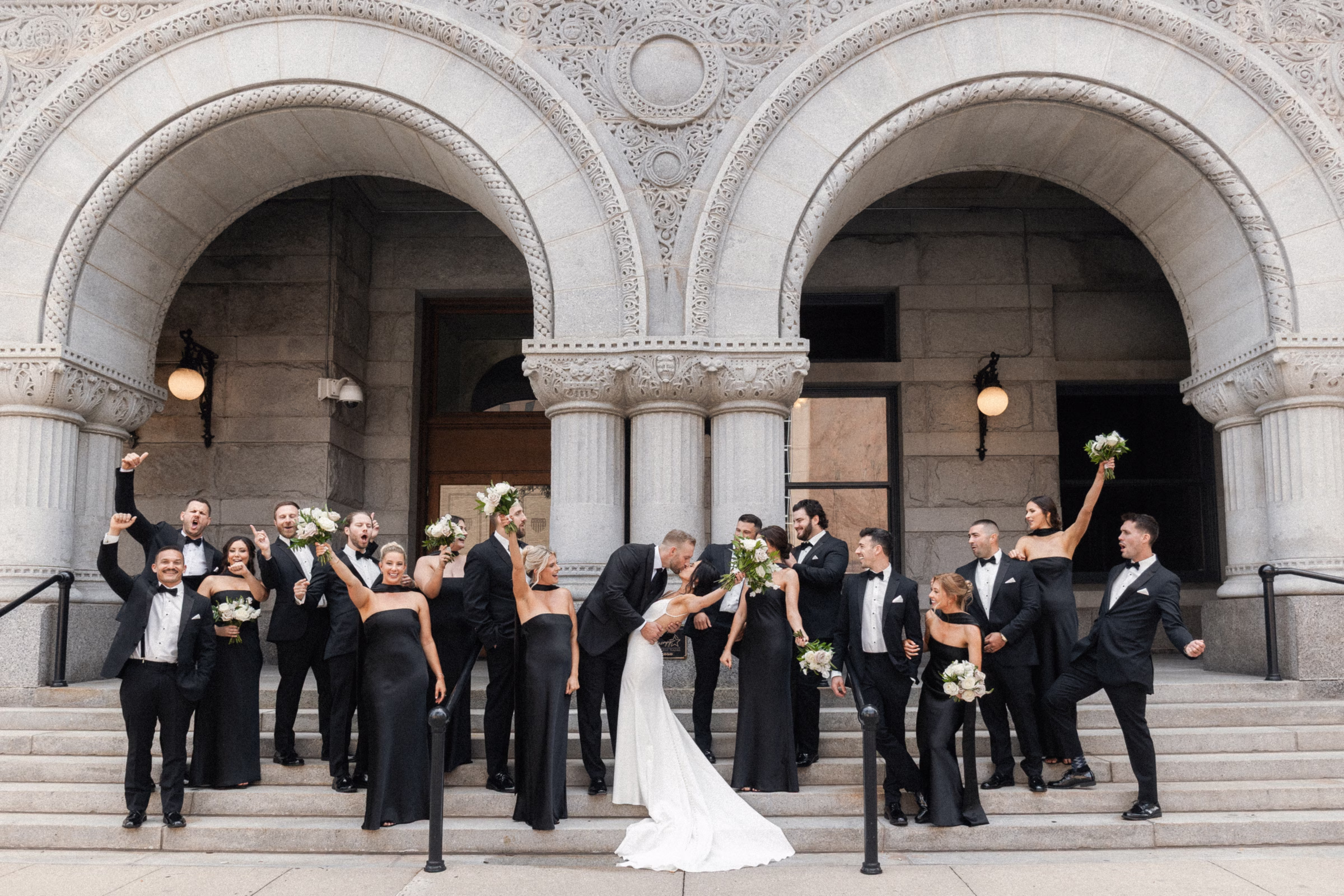 a couple kisses, surrounded by their cheering wedding party