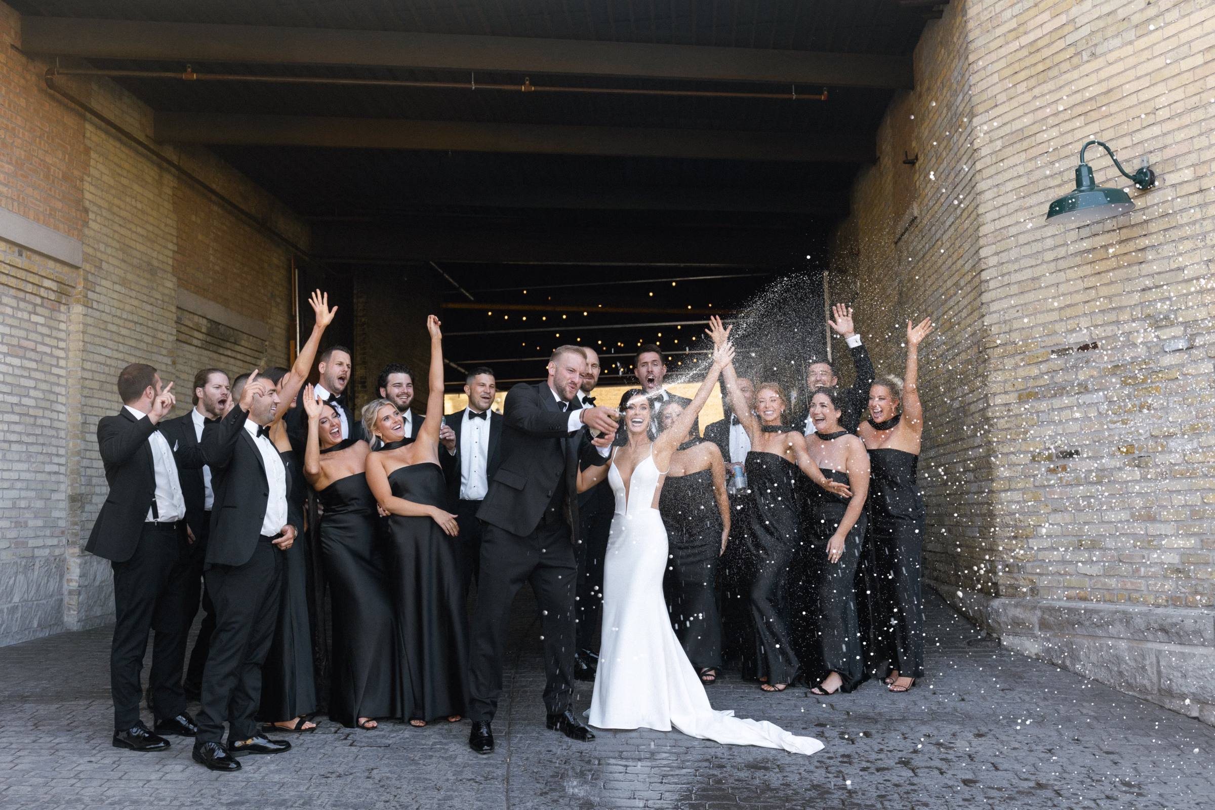 the couple opens a champagne bottle, surrounded by their cheering wedding party