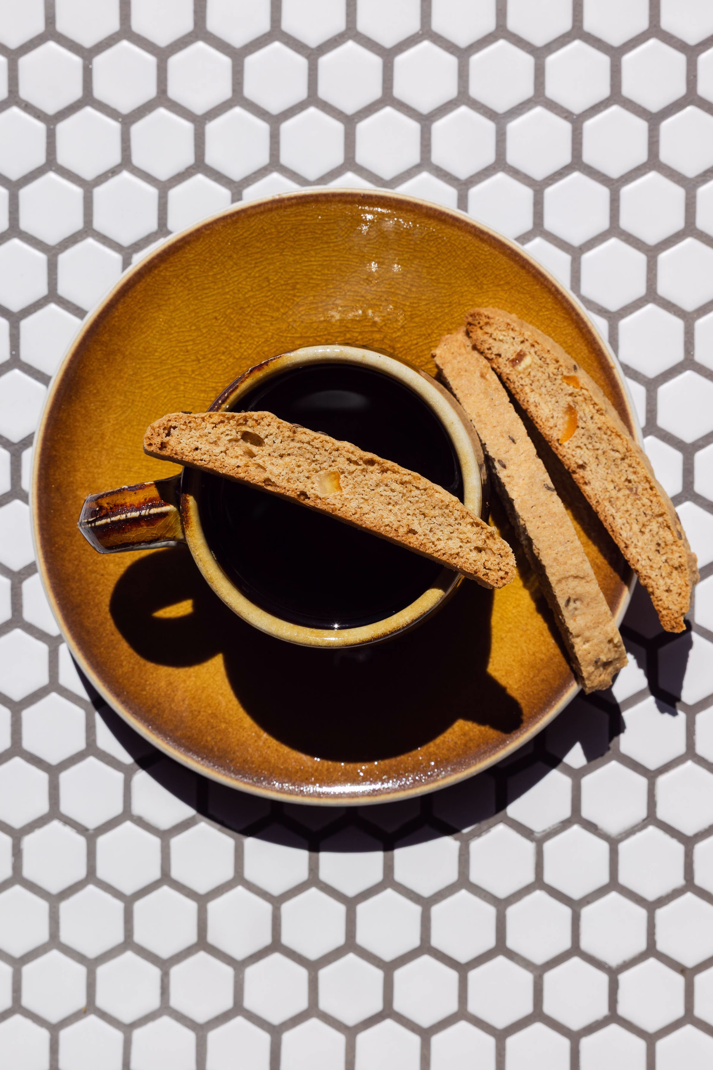 Amati Biscotti biscotti served alongside a cup of coffee on a hexagon tile background