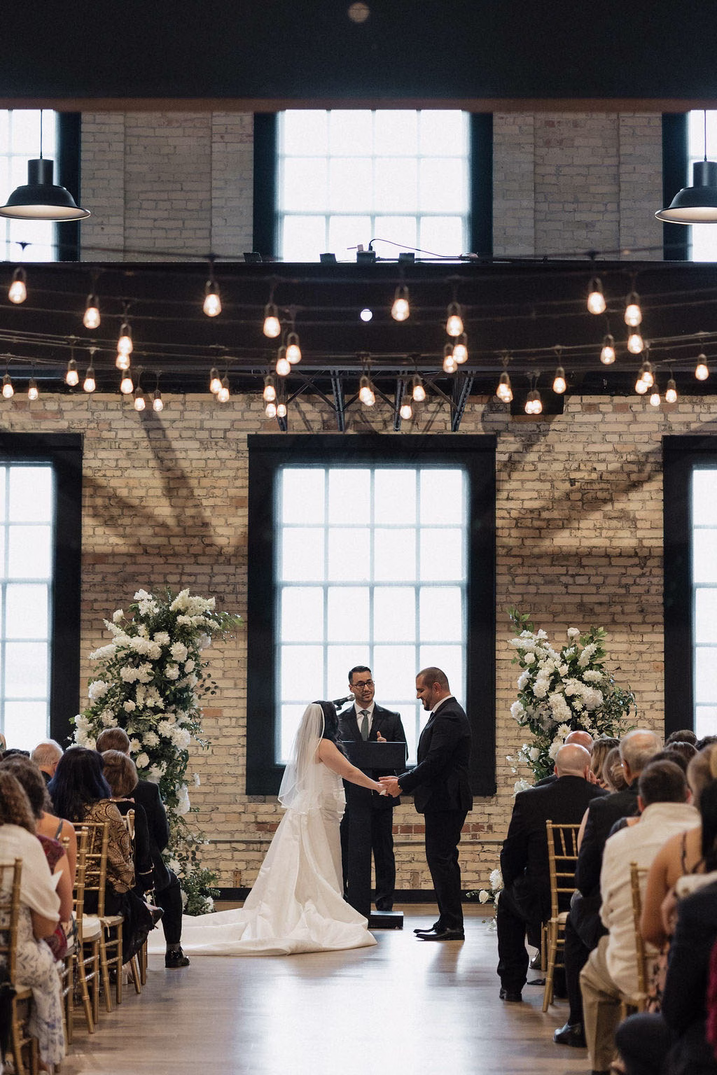 The Berkshire wedding ceremony — couple exchanging vows under exposed brick and string lights with white floral arrangements