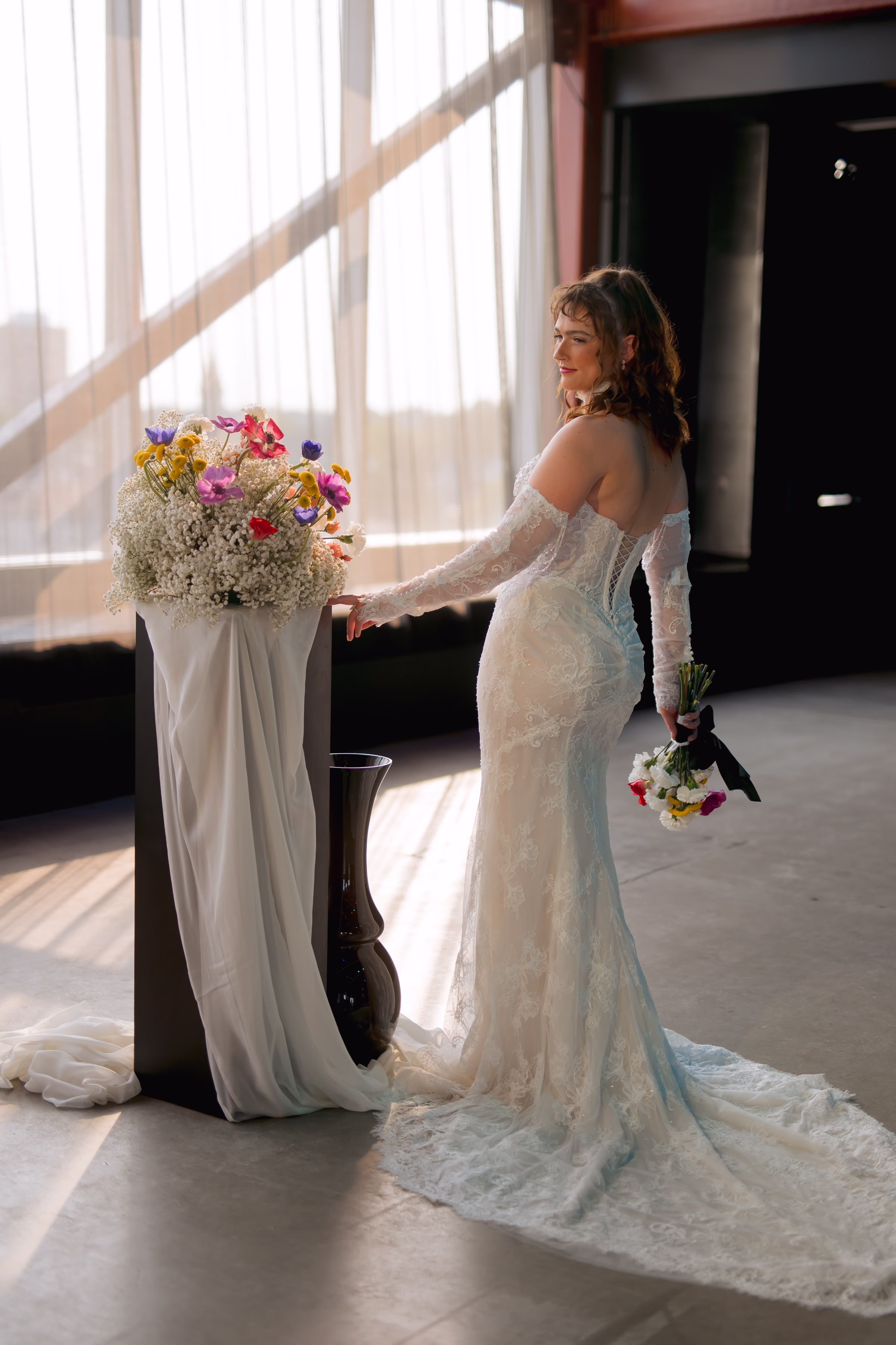 the bride model poses at the ceremony, with one hand placed on a ceremony arch covered in white draping and florals from Flowers for Dreams