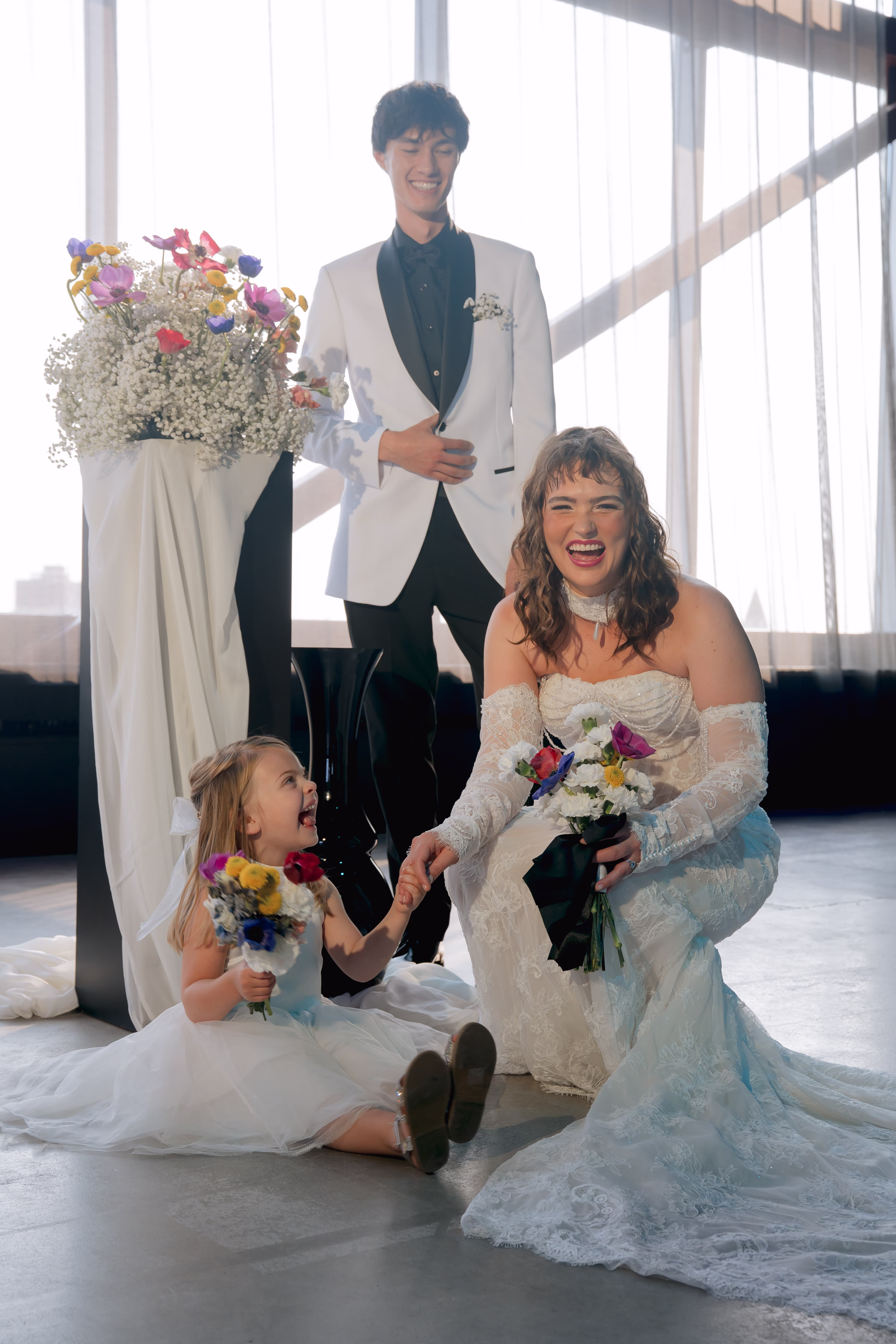 The couple laughs at a wedding ceremony, joined by a flower girl in a white dress