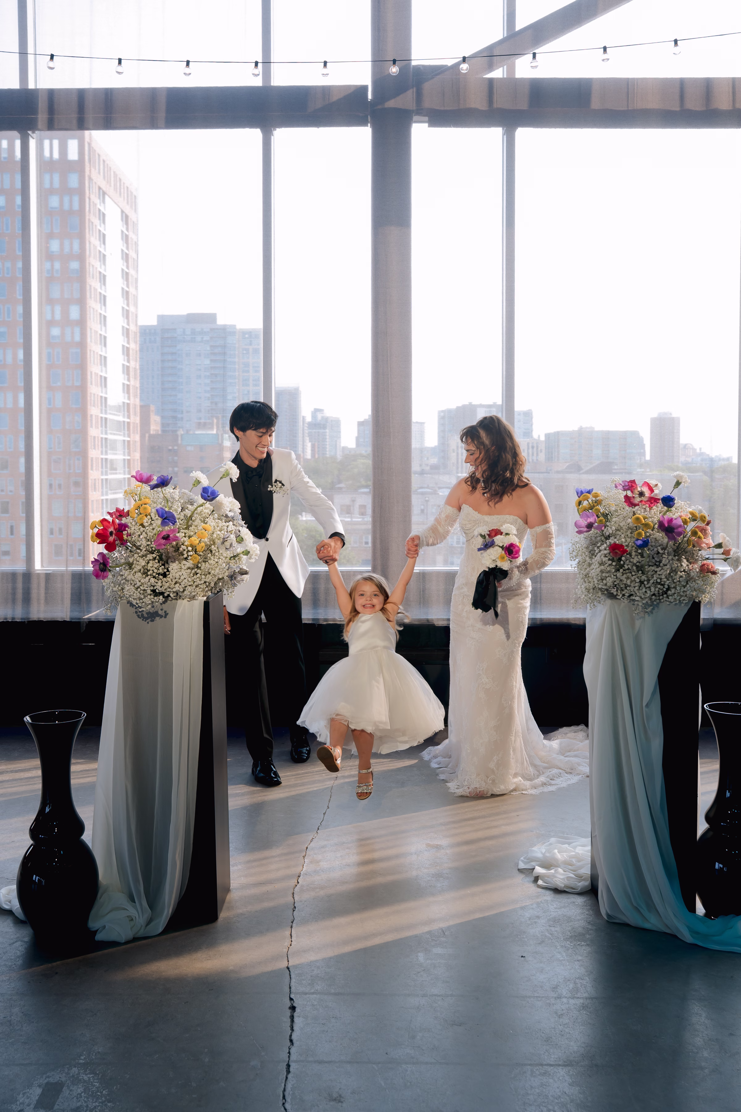 the bride and groom models walk towards the camera at the ceremony set up at Jan Serr Studio, each holding one hand of a young flower girl