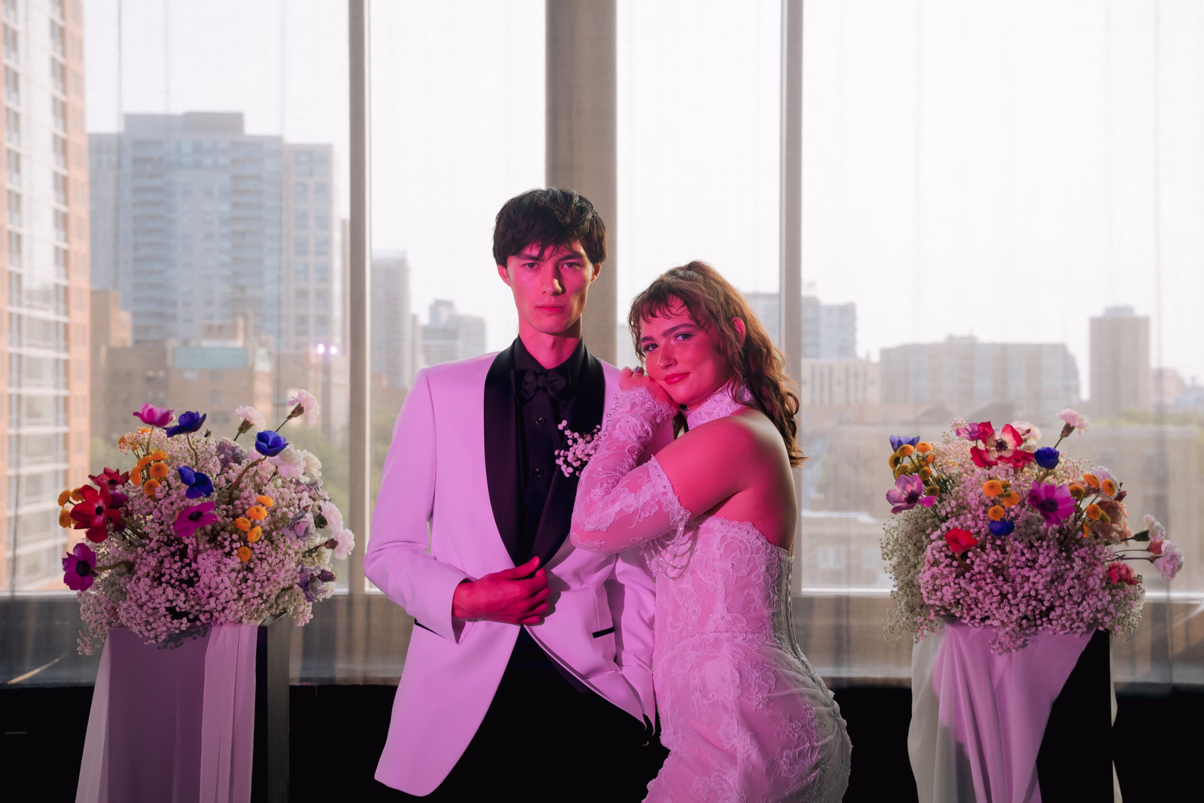 the bride and groom pose at the ceremony space, bathed in pink light, with floral pillars on both sides and views of the Milwaukee skyline visible out the windows behind them.