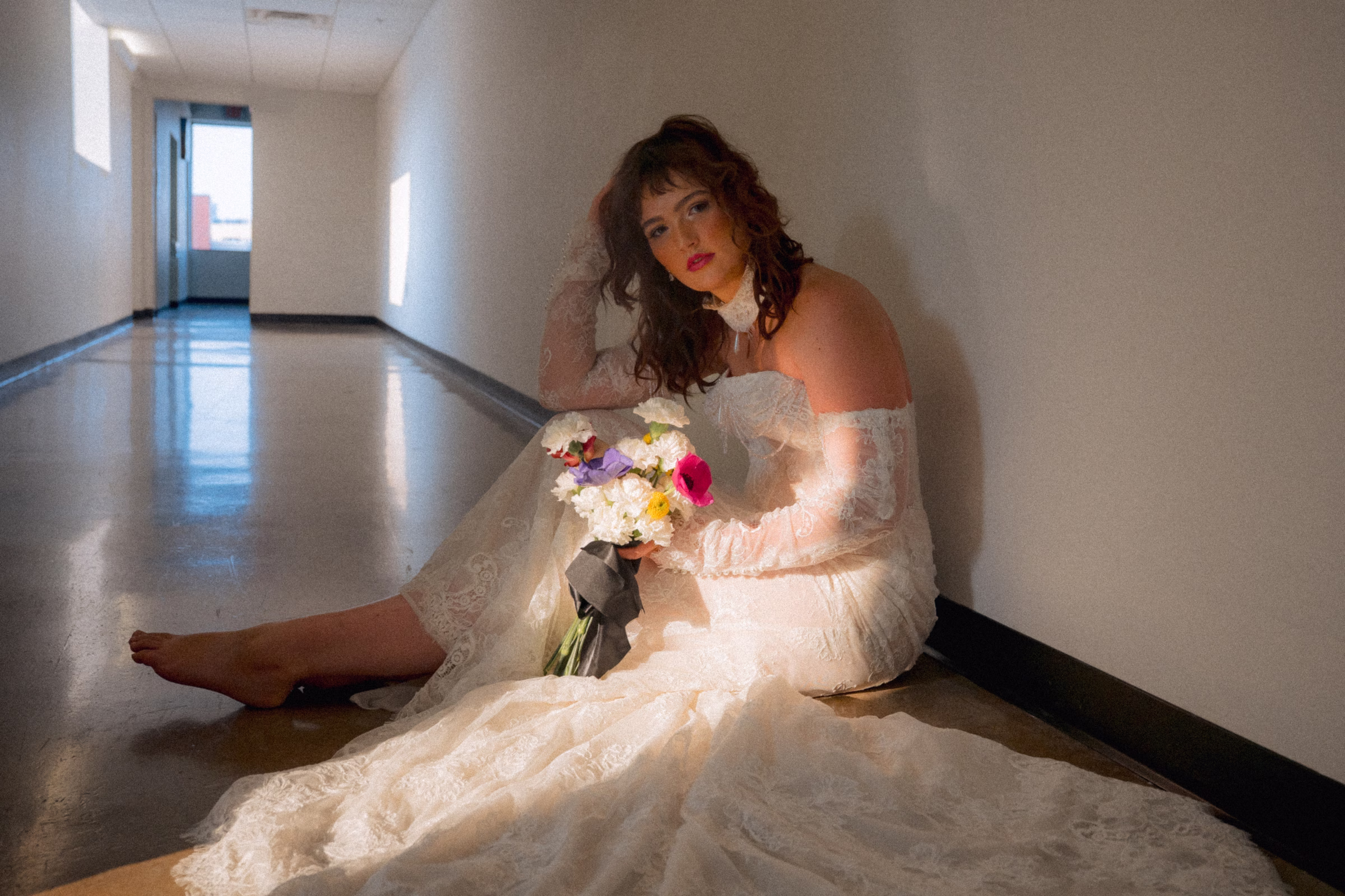 The Rock Agency bridal model, Maggie, poses in the hallway, seated on the floor with light pouring in through the window. She is wearing a delicate lace bridal gown from Miss Ruby Bridal, with lace sleeves, a lace necklace, and delicate train
