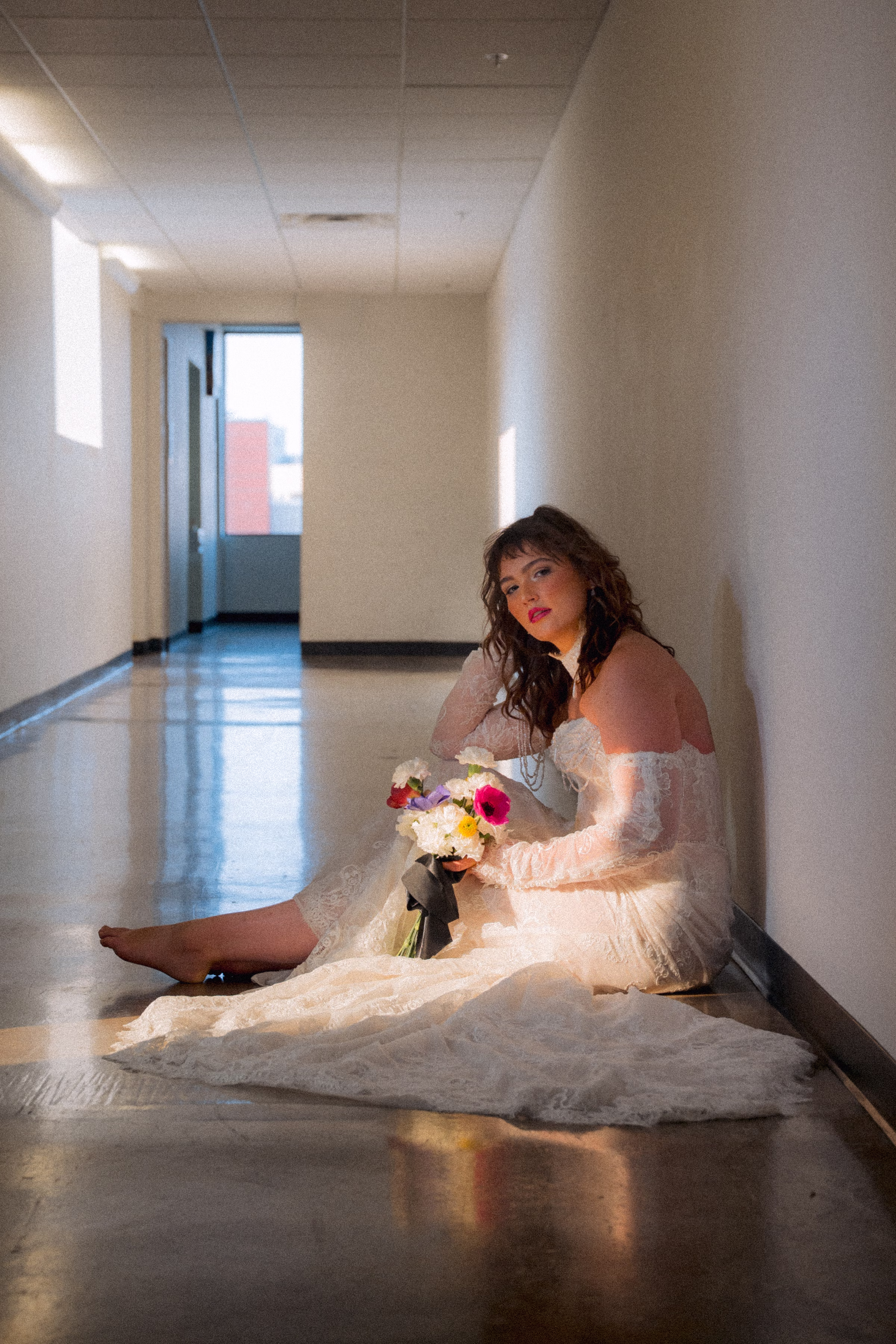 Model Maggie poses in the hallway, seated on the floor with light pouring in through the window. She is wearing a lace bridal gown from Miss Ruby Bridal, with lace sleeves, a lace necklace, and delicate train. She is holding a bouquet from Flowers for Dreams
