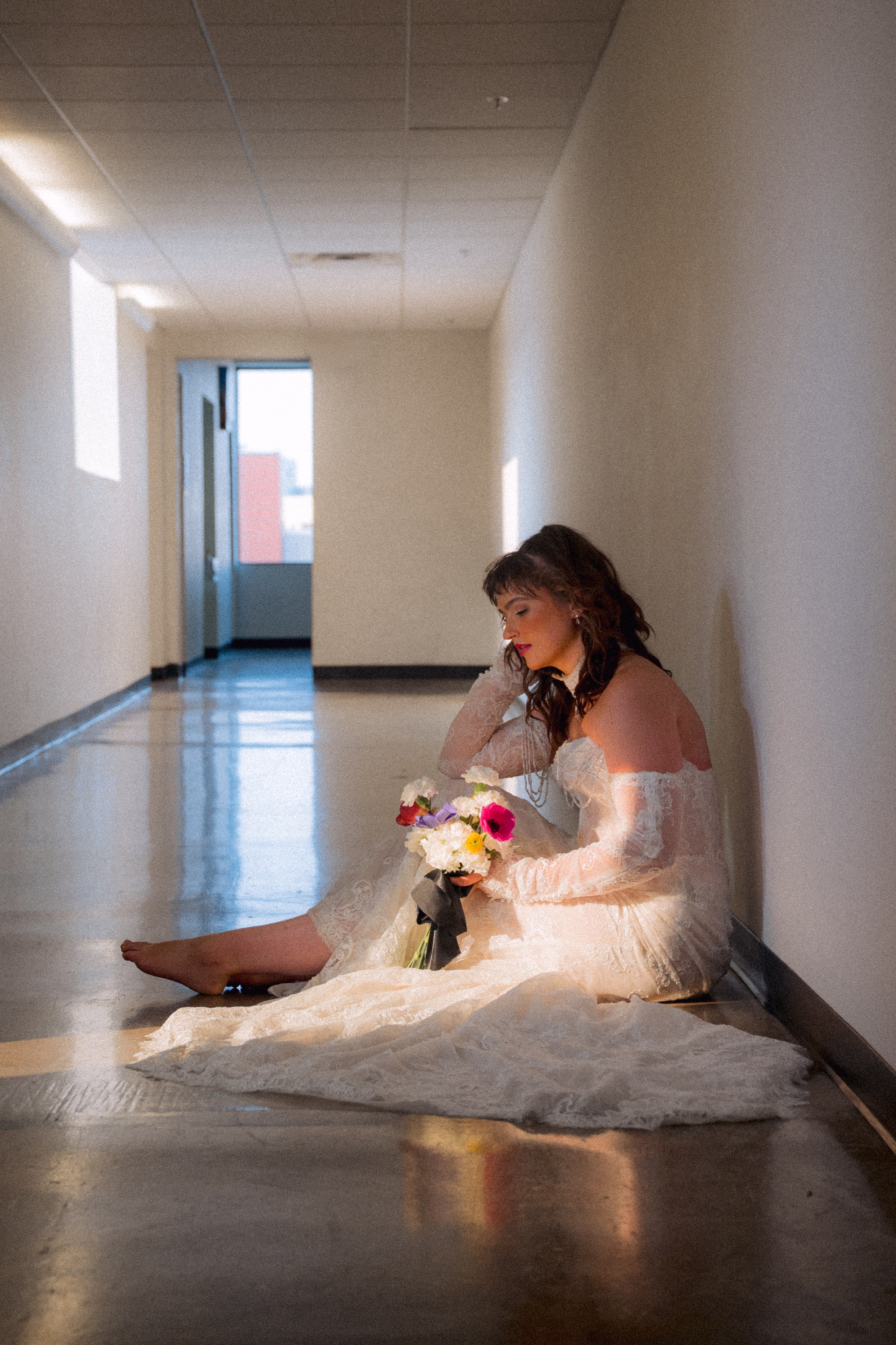 Model Maggie poses in the hallway, seated on the floor with light pouring in through the window. She is wearing a lace bridal gown from Miss Ruby Bridal, with lace sleeves, a lace necklace, beaded details, and delicate train. She is holding a bouquet from Flowers for Dreams