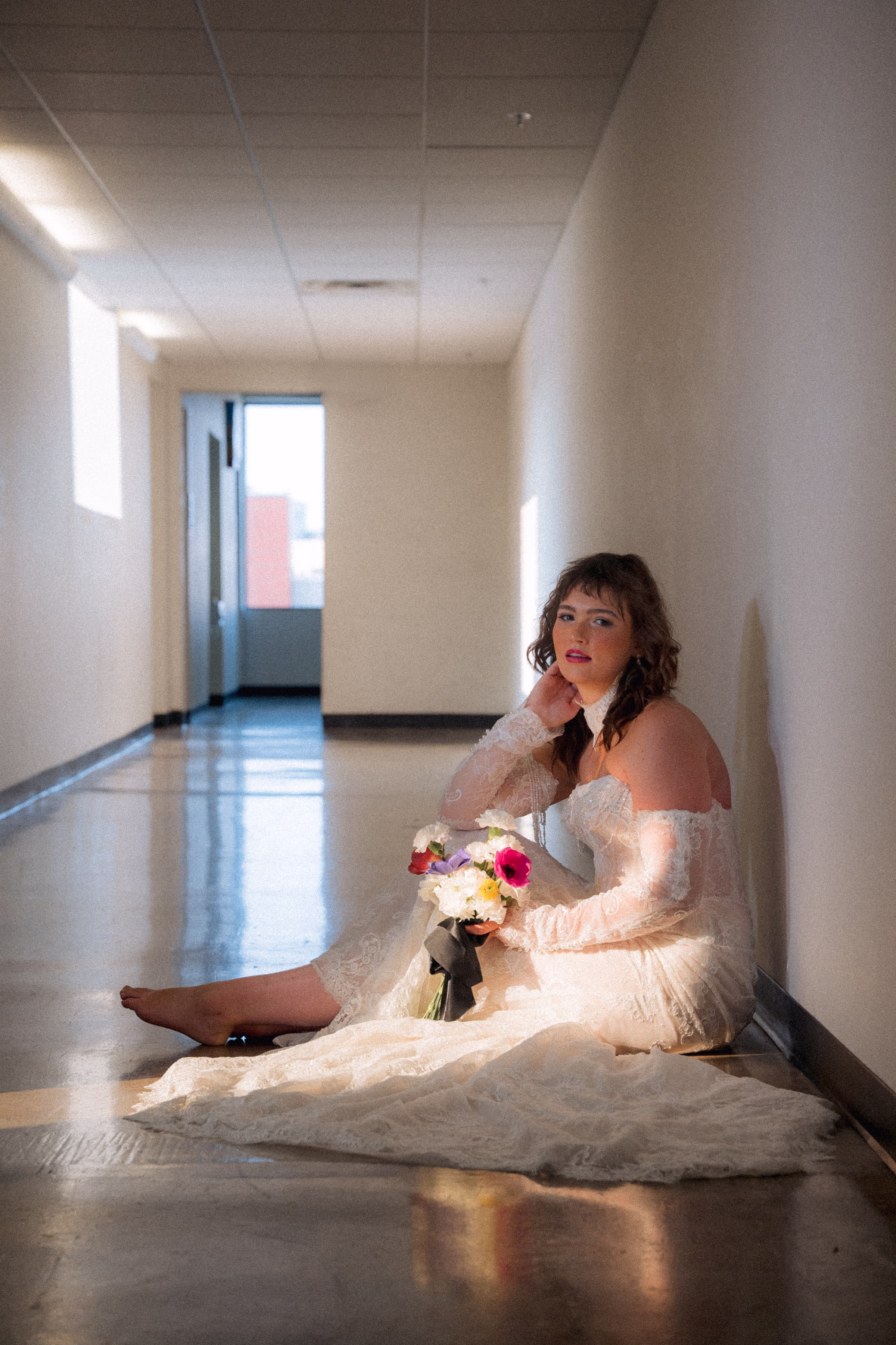 Model Maggie poses in the hallway of Jan Serr Studio, seated on the floor with light pouring in through the window. She is wearing a lace bridal gown from Miss Ruby Bridal, with lace sleeves, a lace necklace, and delicate train. She is holding a bouquet from Flowers for Dreams