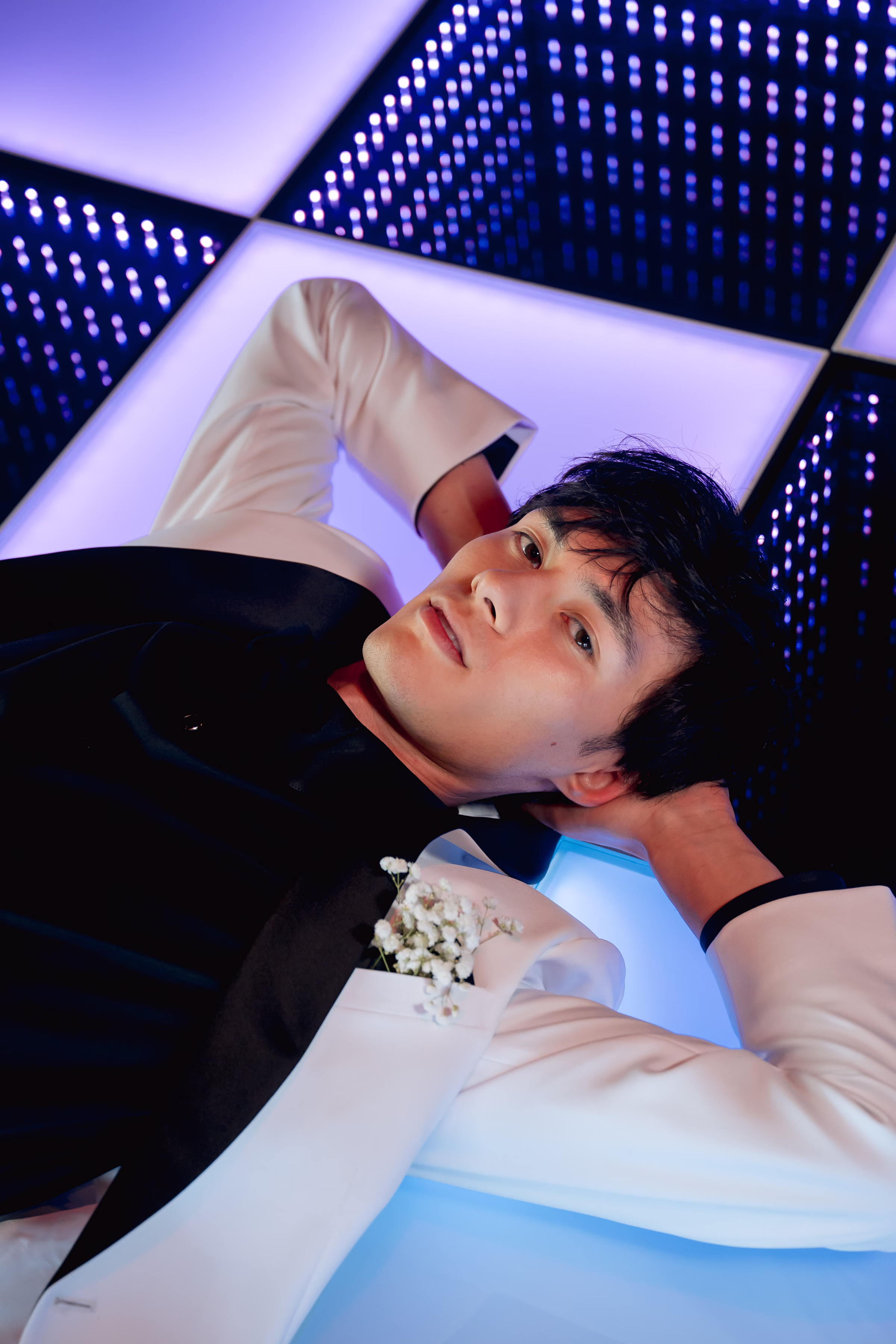a close-up photo of a groom lounging on an LED dance floor. He has a baby's breath boutonniere pinned to the lapel of his suit jacket from DuBois Formalwear