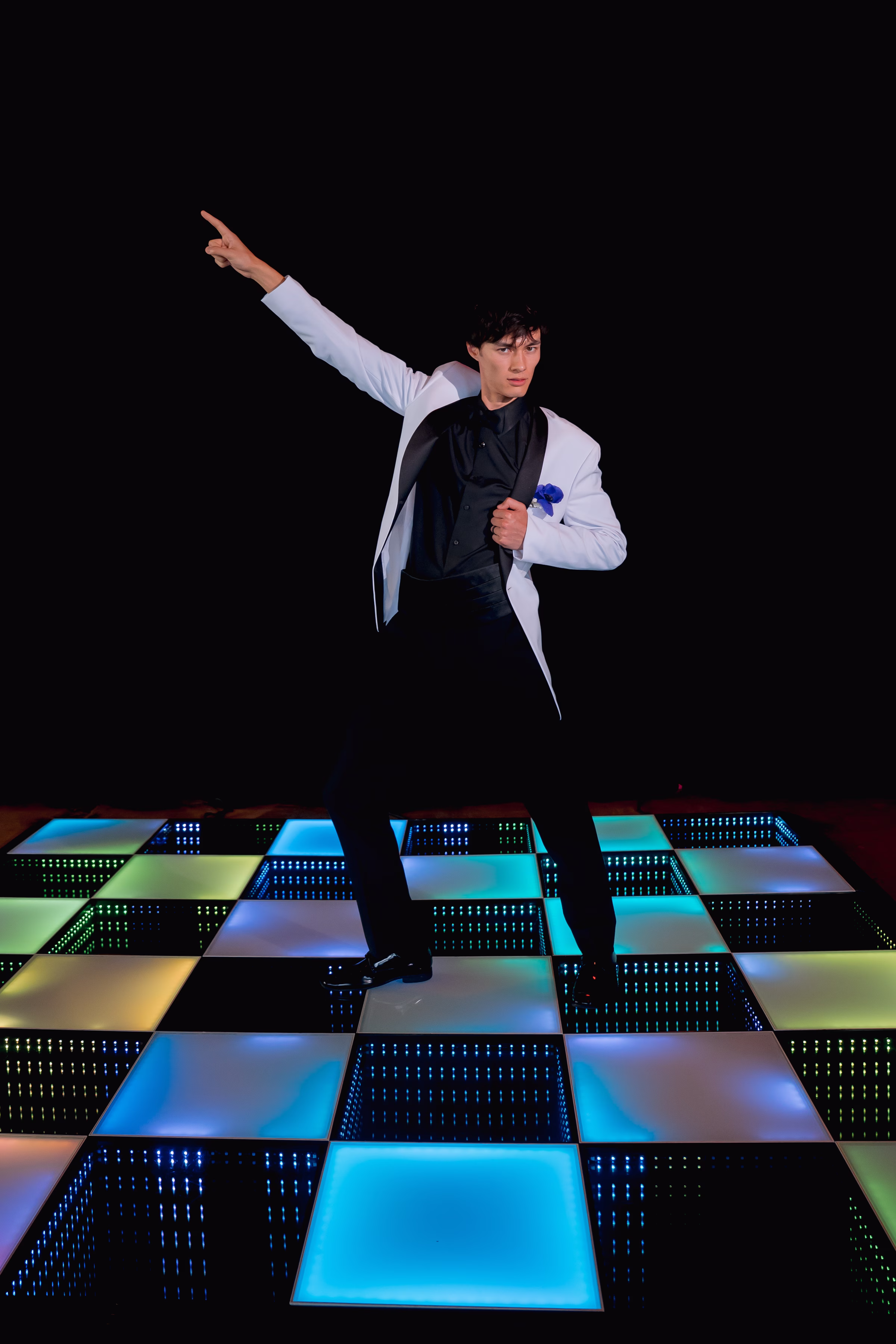 a groom model does a disco dance on a colorful LED dance floor, with one hand pointing and the other on the lapel of his white suit jacket from DuBois Formalwear