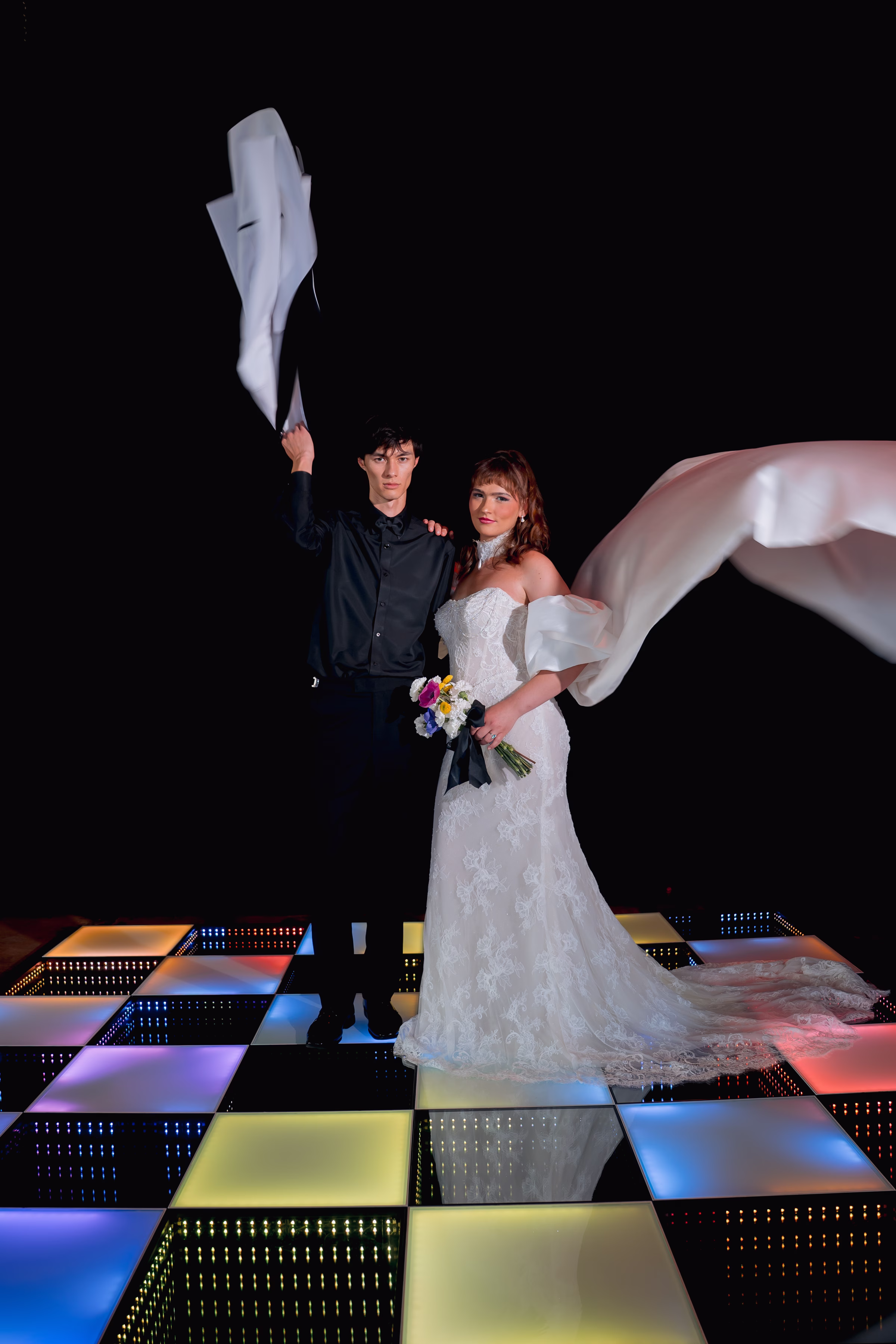 a bride and groom post on a colorful LED dance floor. The groom is swinging around his white suit jacket, and the bride's reception cape is flowing behind her