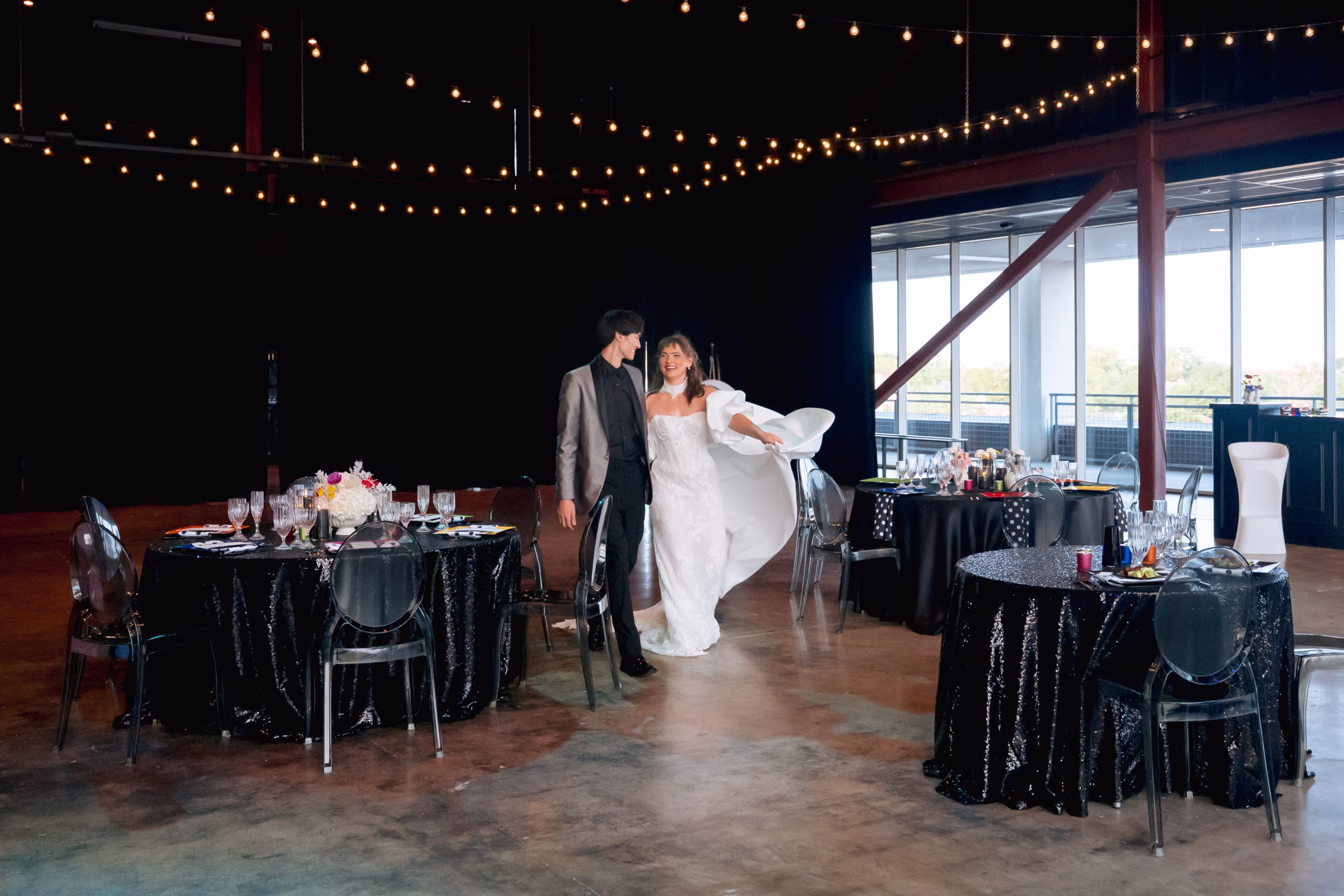 the bride and groom smile at each other while walking through the bold wedding inspiration reception space at Jan Serr Studio. They are showcasing a second fashion look from Miss Ruby Bridal, with the bride wearing puffy off-the-shoulder sleeves and a cape, while the groom is now wearing a silver suit jacket