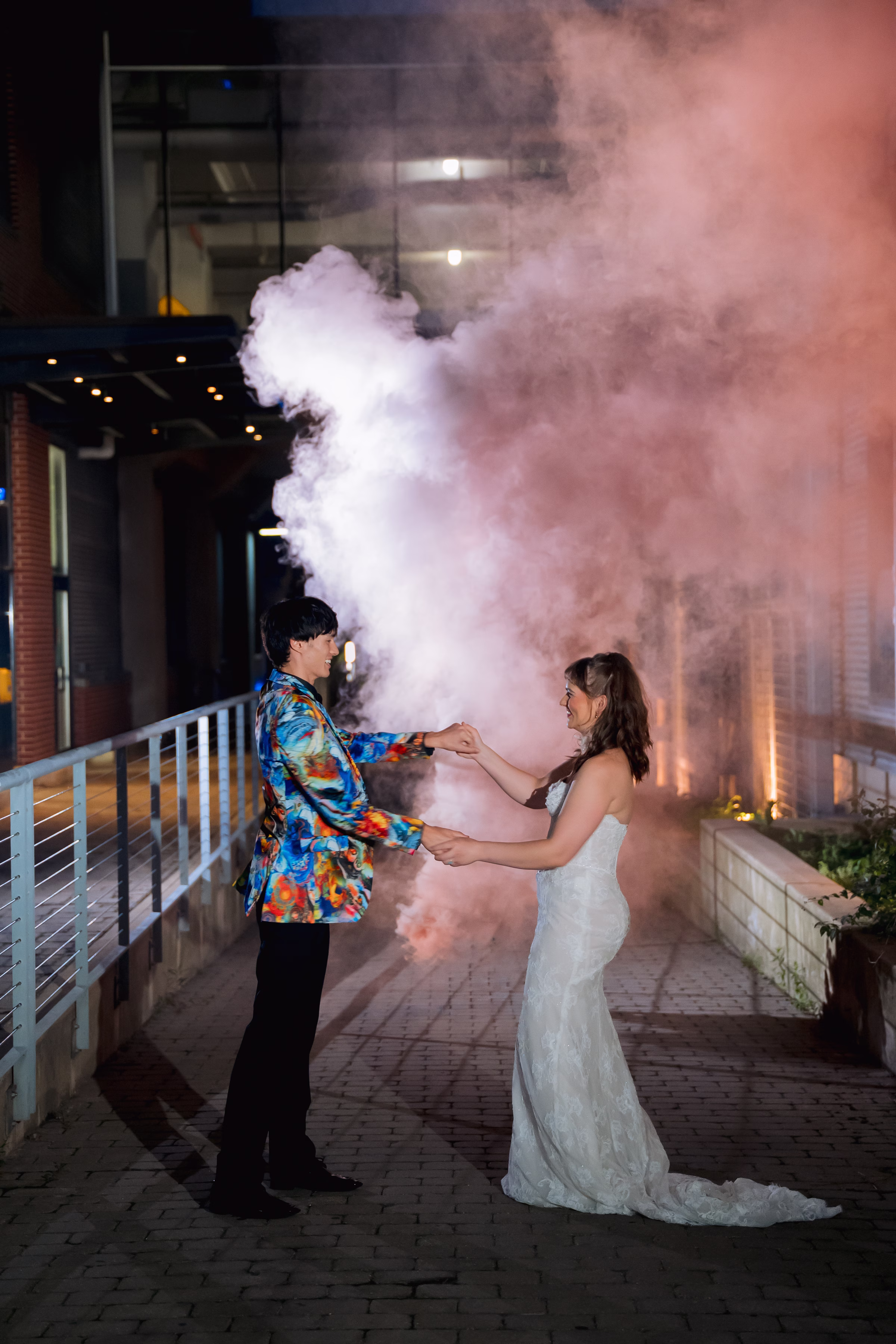 a bride and groom pose for dramatic, urban photos with smoke bombs from Flowers for Dreams going off behind them. The bride is wearing a dress from MIss Ruby Bridal, and the groom is wearing all black, aside from a colorful suit-jacket from DuBois Formalwear