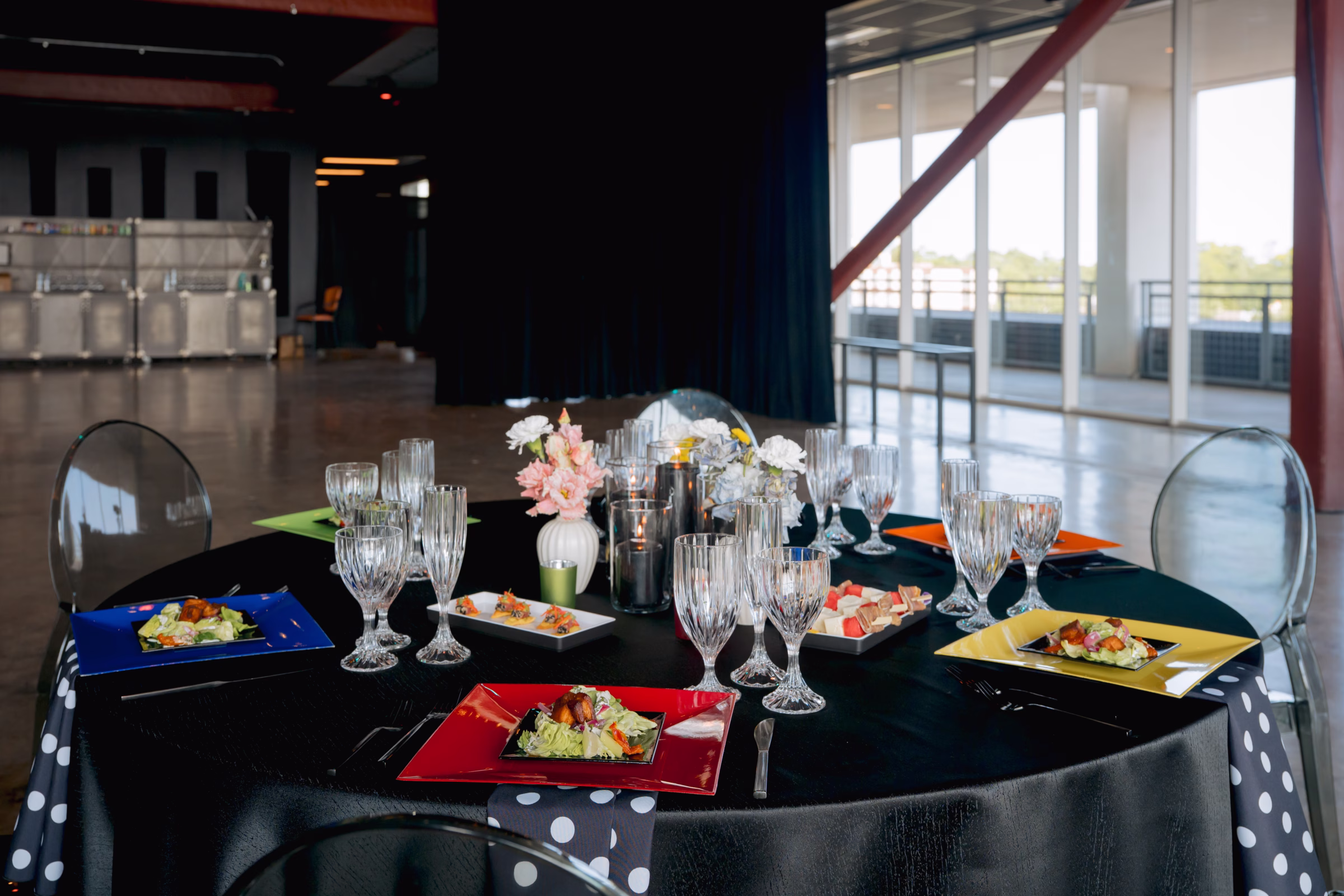 a tablescape featuring rentals from Windy City Linens and Tablescape Event Rentals. Boldly colored square plates in red, yellow, blue, and orange sit a round table with a black tablecloth, crystal glassware, floral centerpieces, and black-and-white polka dot napkins