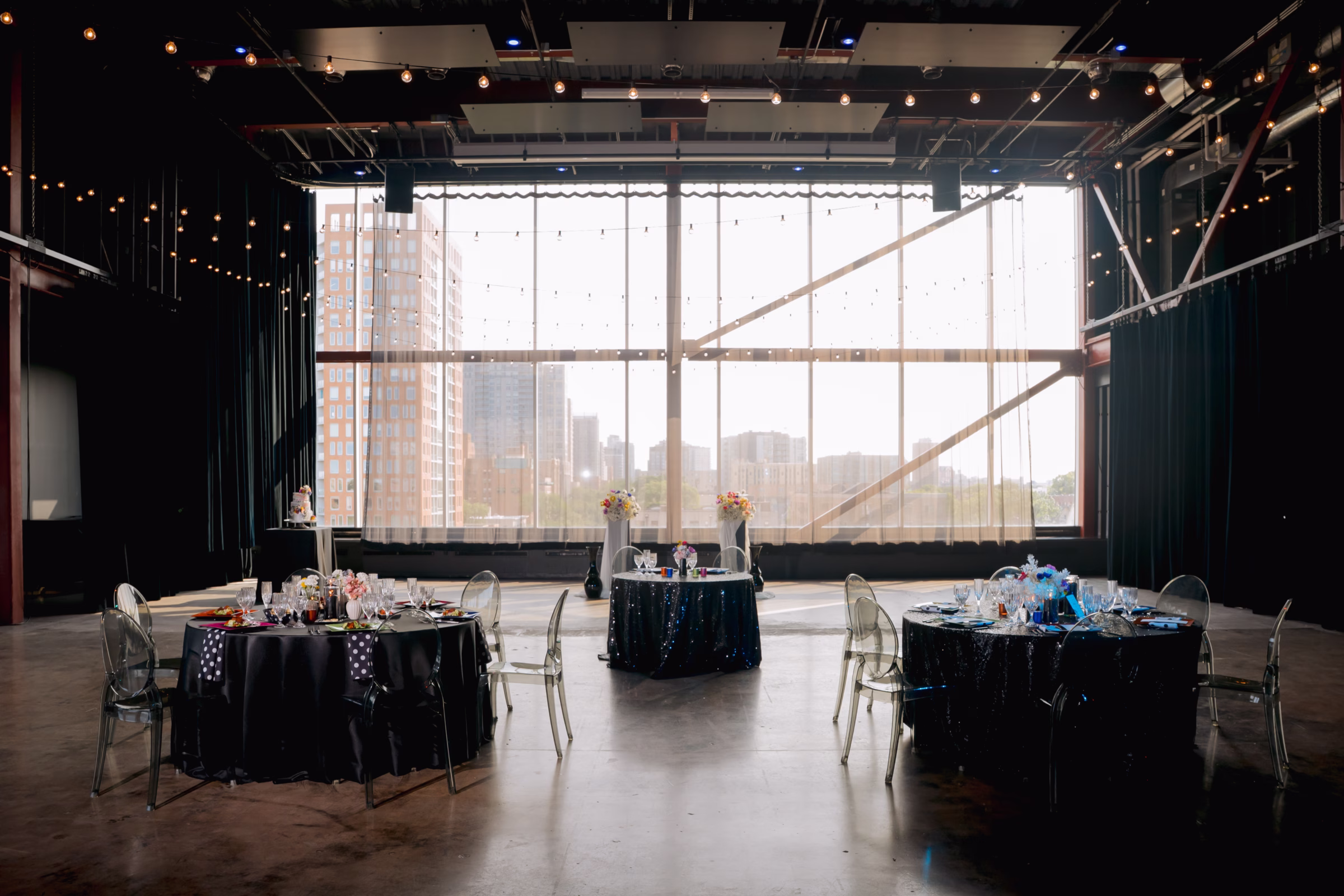 a landscape photo showing industrial meets modern events space, Jan Serr Studio, located on the top floor of UWM's Kenilworth Square East building. The venue has floor-to-ceiling windows and panoramic views of Milwaukee's Downtown Skyline. There are three reception tables and the ceremony space visible in the background