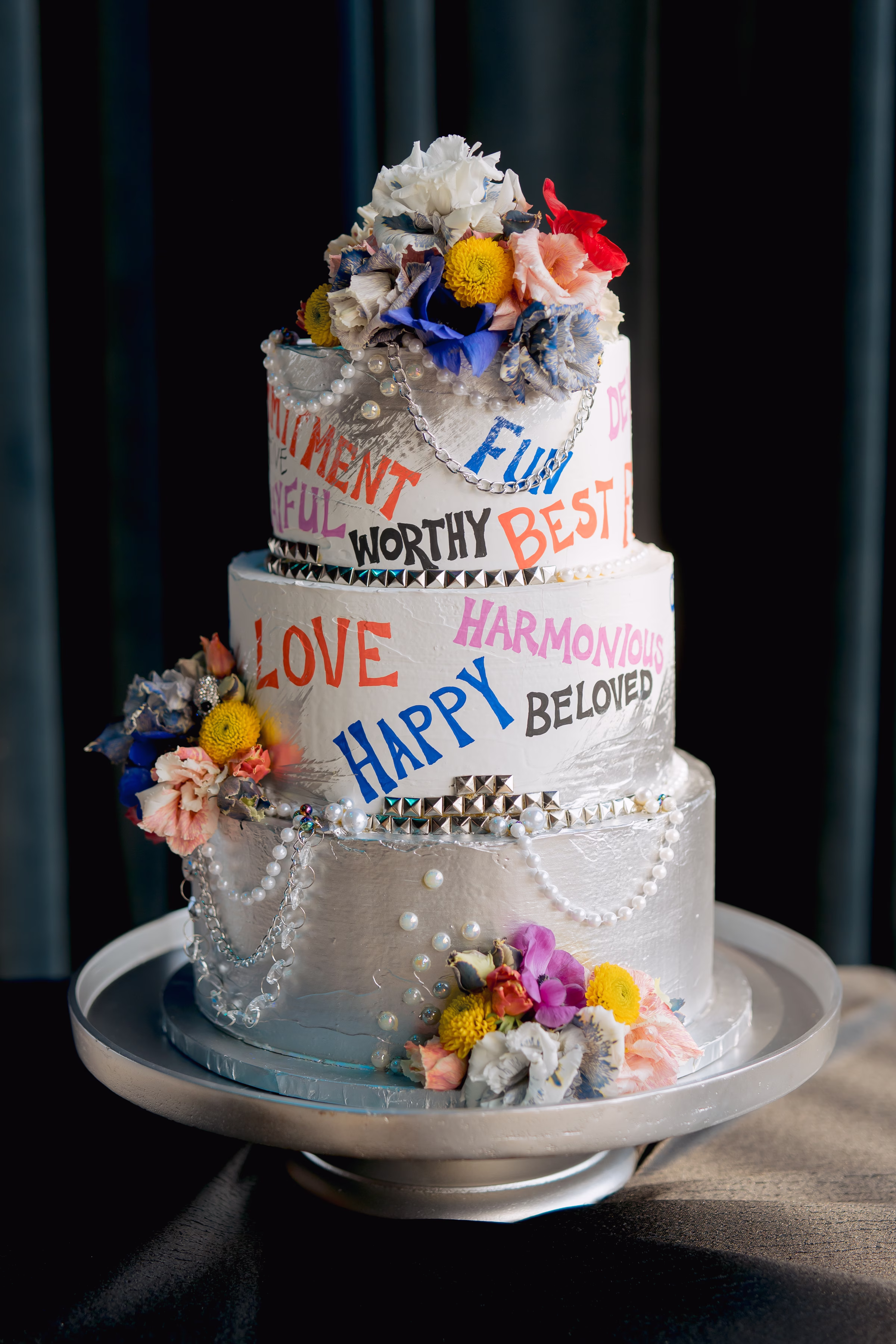 a three-tier, graffiti-inspired cake design by Cowboy David's Bakery featuring a silver base layer with pearl details, and colorful words of affirmation written on the middle and top layers