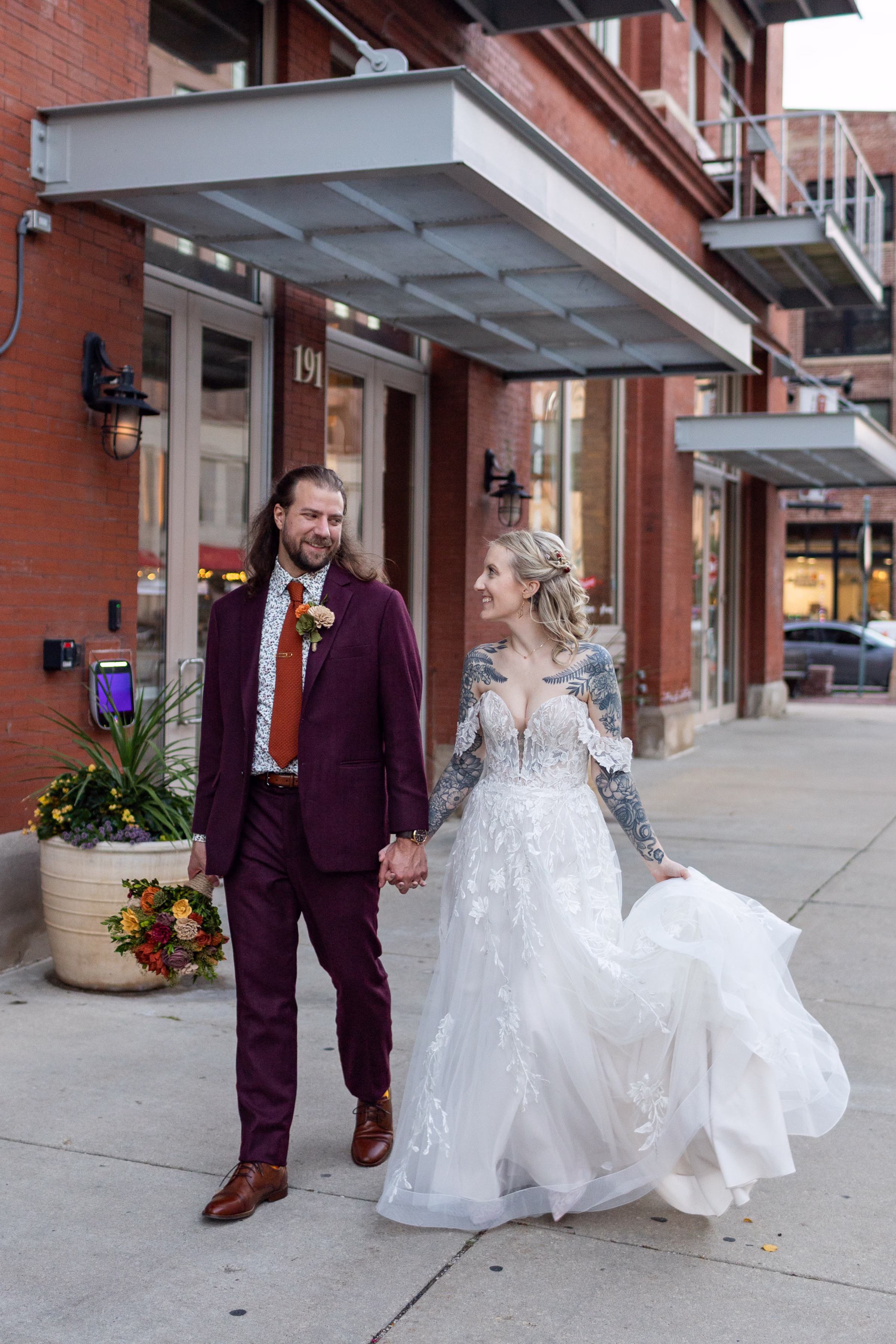 moody romantic fall wedding loft on broadway milwaukee couple portraits 654 11508 - A Moody, Romantic Fall Wedding at The Loft on Broadway | Sarah + Nathan a bride and groom hold hands and walk down the street in Milwaukee, Wisconsin. The bride is wearing an ornate, delicate lace dress from Strike Bridal Bar with illusion deep-V neckline, off-the-shoulder sleeves, and a layered tulle skirt