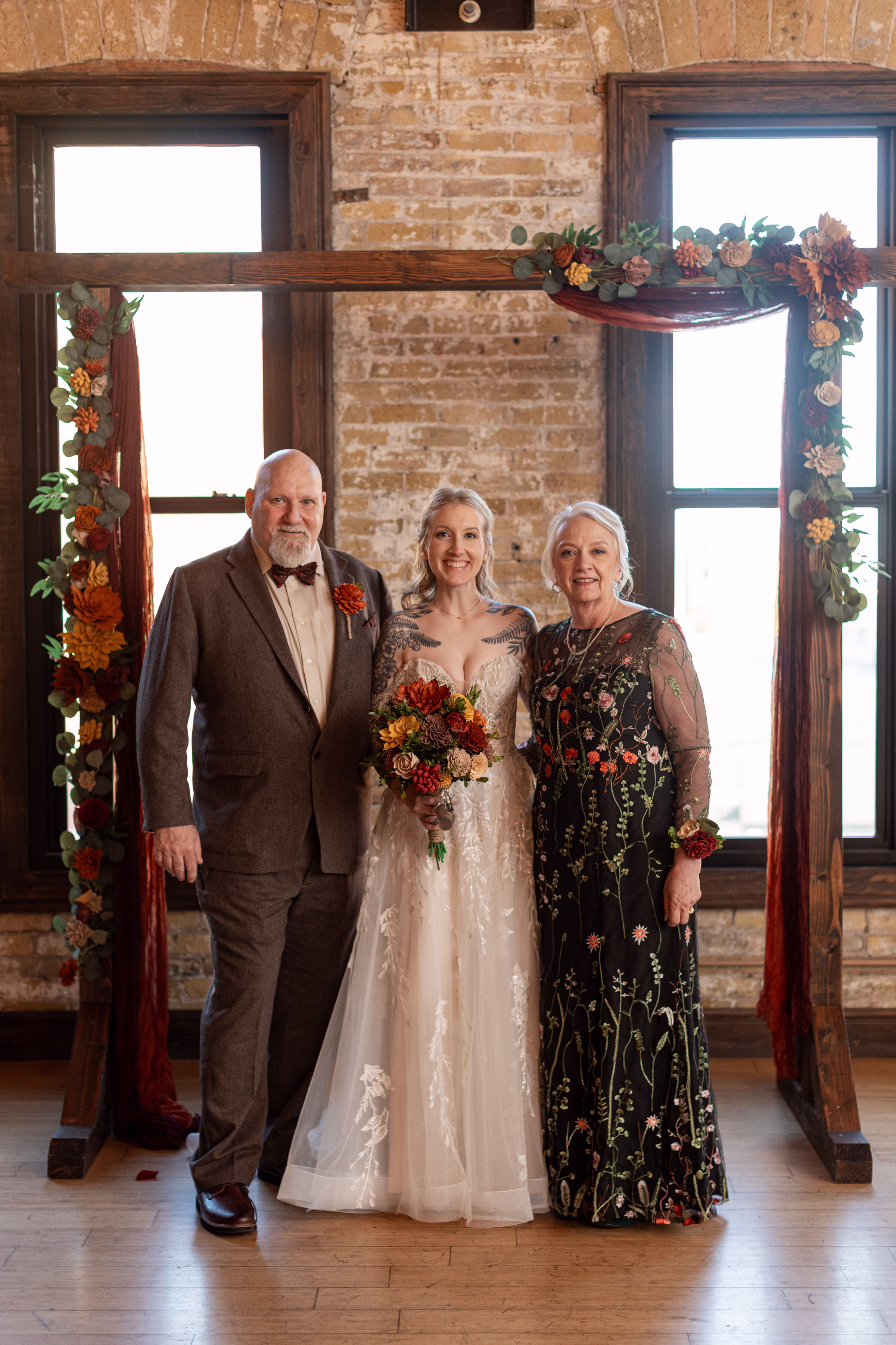 moody romantic fall wedding loft on broadway milwaukee group portraits 493 0935 - A Moody, Romantic Fall Wedding at The Loft on Broadway | Sarah + Nathan a bride poses with her parents on either side in front of a wooden ceremony backdrop
