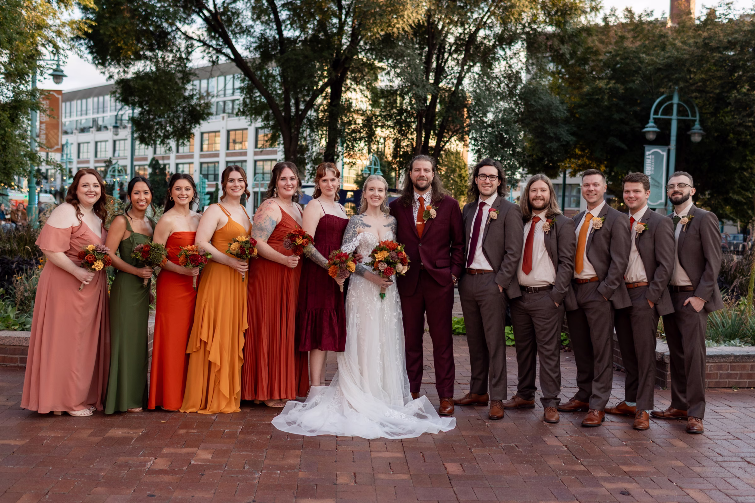 moody romantic fall wedding loft on broadway milwaukee group portraits 515 10999 - A Moody, Romantic Fall Wedding at The Loft on Broadway | Sarah + Nathan Sarah & Nathan stand in a line with their wedding party, with bridesmaids on the left and groomsmen on the right. The groomsmen's ties coordinate with the varying colors of bridesmaids dresses in burgundy, burnt orange, tangerine, red, green, and mauve