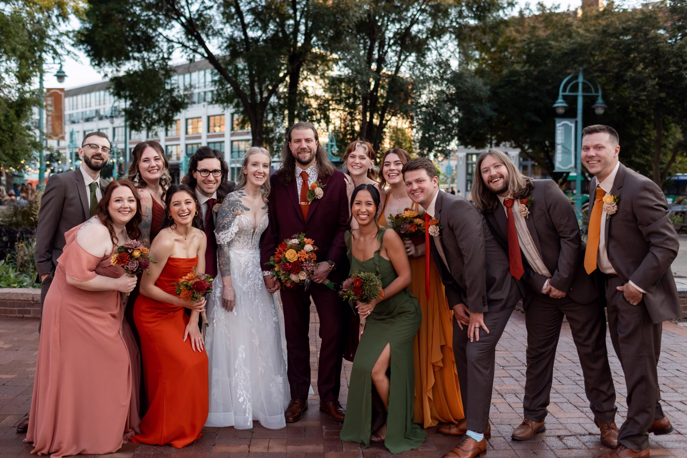 moody romantic fall wedding loft on broadway milwaukee group portraits 545 11101 - A Moody, Romantic Fall Wedding at The Loft on Broadway | Sarah + Nathan a bride and groom smile alongside their wedding party, dressed in a moody fall color palette, as the sun begins to set behind them