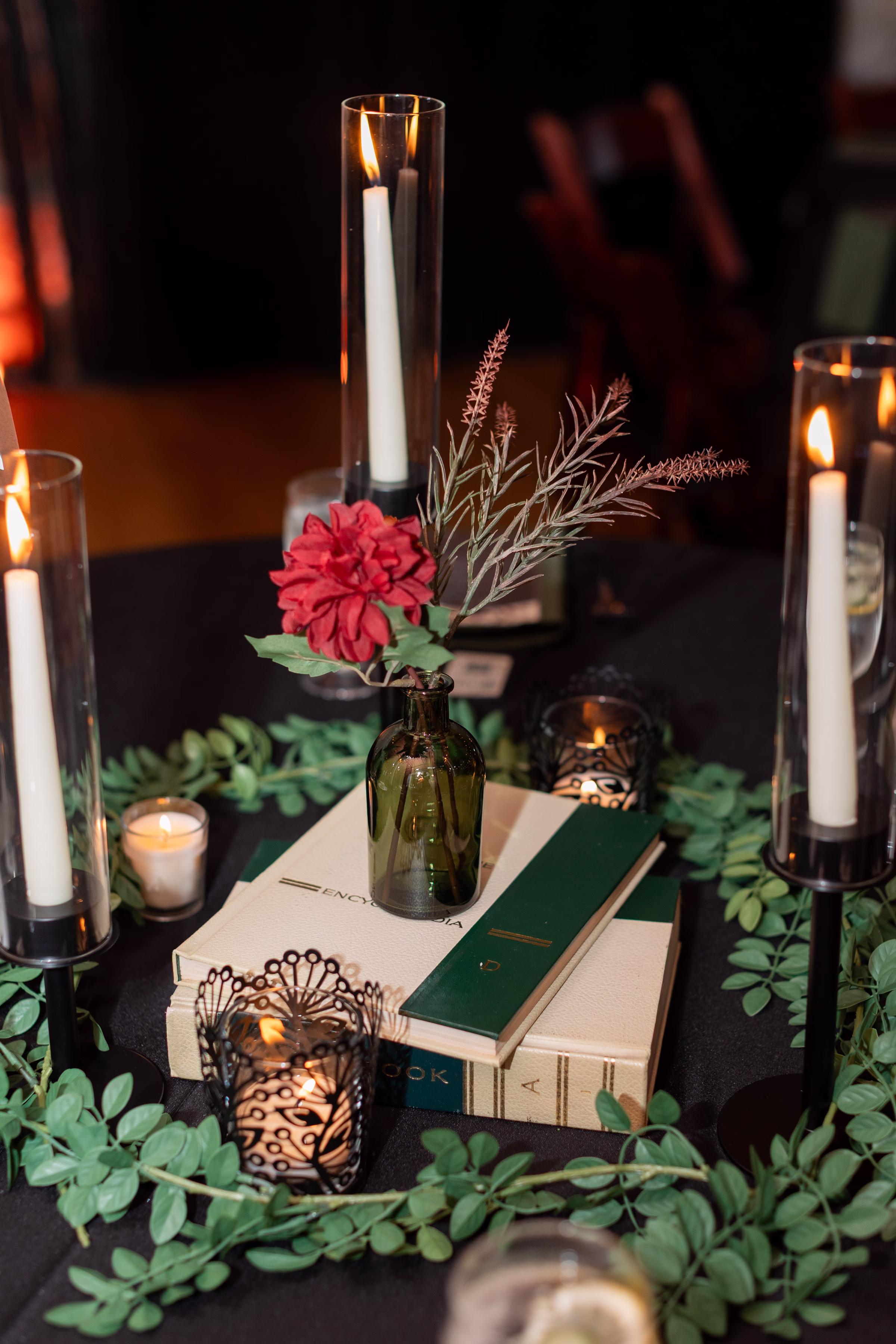 moody romantic fall wedding loft on broadway milwaukee reception centerpieces table numbers tarot 675 11515 - A Moody, Romantic Fall Wedding at The Loft on Broadway | Sarah + Nathan a close-up on a wedding reception table centerpiece consisting of stacked books, a bud vase, greenery, and lit candles of varying heights