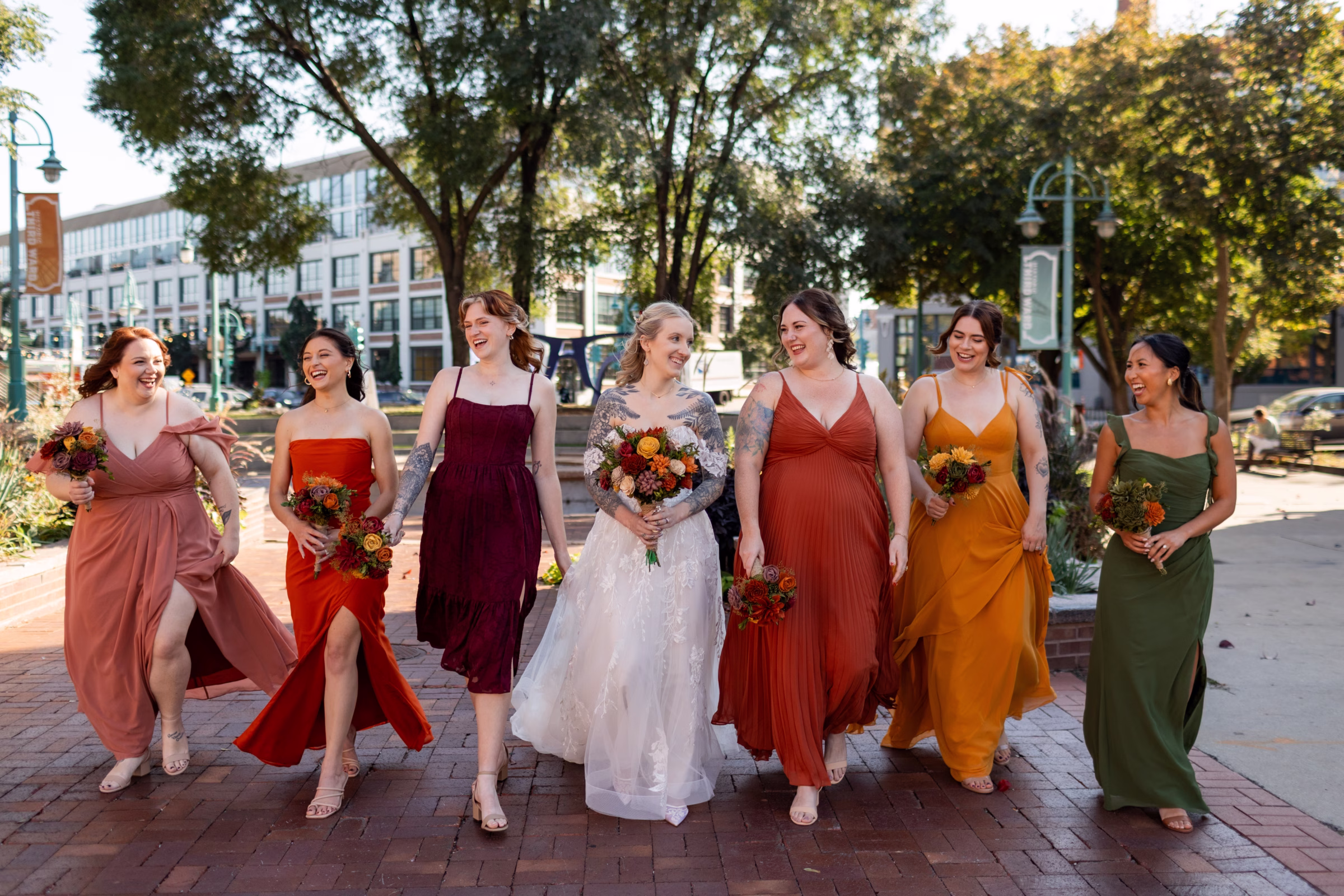 moody romantic fall wedding loft on broadway milwaukee wisconsin bridesmaids portraits 237 0476 - A Moody, Romantic Fall Wedding at The Loft on Broadway | Sarah + Nathan a bride walks alongside six bridesmaids in colorful fall-palette dresses. All are holding wood flower bouquets