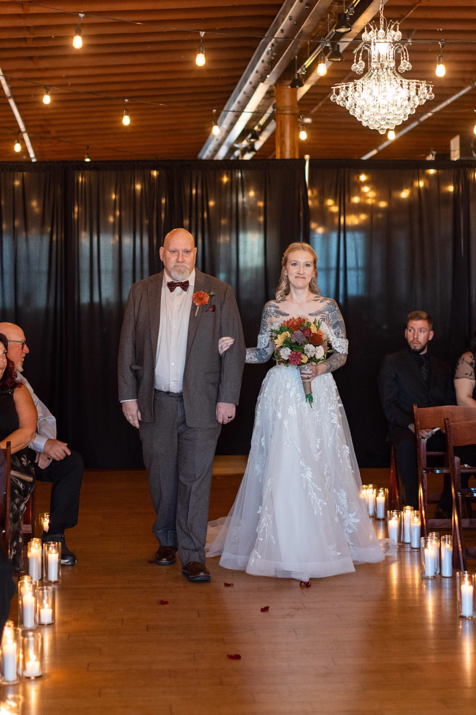 moody romantic fall wedding loft on broadway milwaukee wisconsin ceremony 436 0804 - A Moody, Romantic Fall Wedding at The Loft on Broadway | Sarah + Nathan a bride is walked down a candle-lit aisle by her father at The Loft on Broadway in Milwaukee, Wisconsin. There is black drapery behind her, along with glowing lights and chandeliers reflecting off the wooden floors and ceiling
