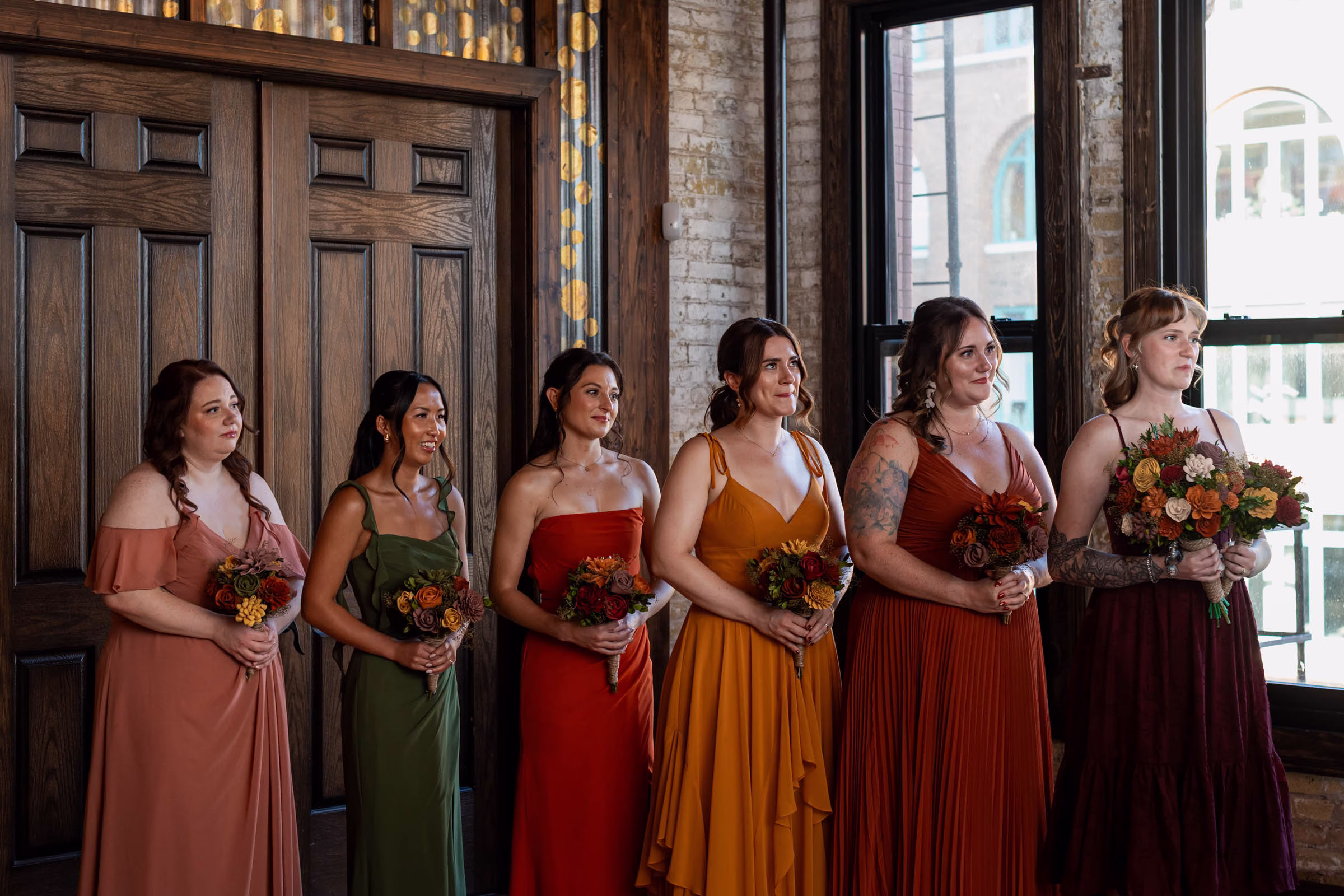 moody romantic fall wedding loft on broadway milwaukee wisconsin ceremony 454 0826 - A Moody, Romantic Fall Wedding at The Loft on Broadway | Sarah + Nathan bridesmaids wearing dresses in varying fall colors stand at the end of one side of the aisle