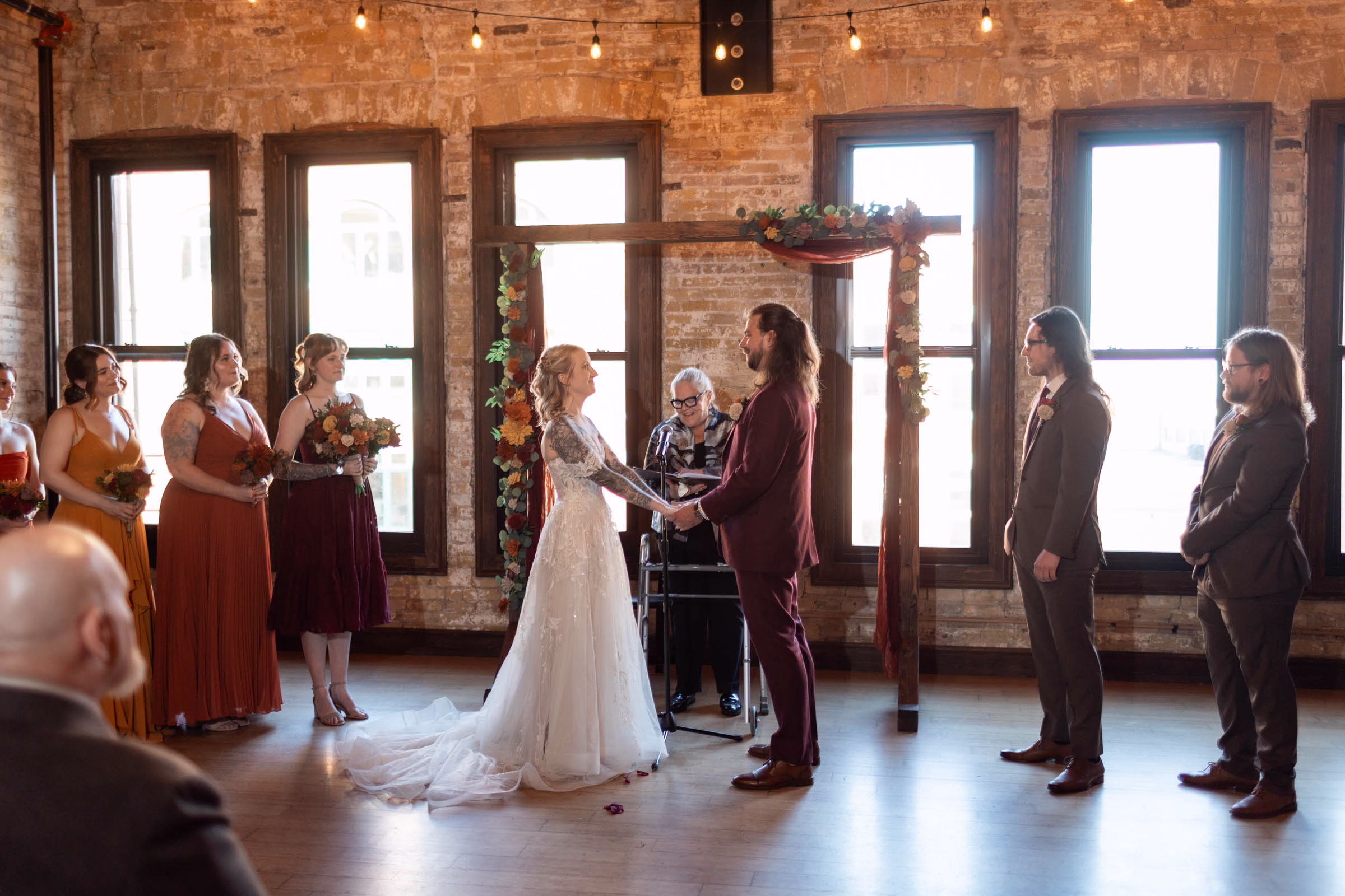 moody romantic fall wedding loft on broadway milwaukee wisconsin ceremony 455 10857 - A Moody, Romantic Fall Wedding at The Loft on Broadway | Sarah + Nathan Sarah & Nathan hold hands at their moody, romantic fall wedding at The Loft on Broadway. There is a wooden backdrop behind them with colorful flowers and burgundy tulle draping